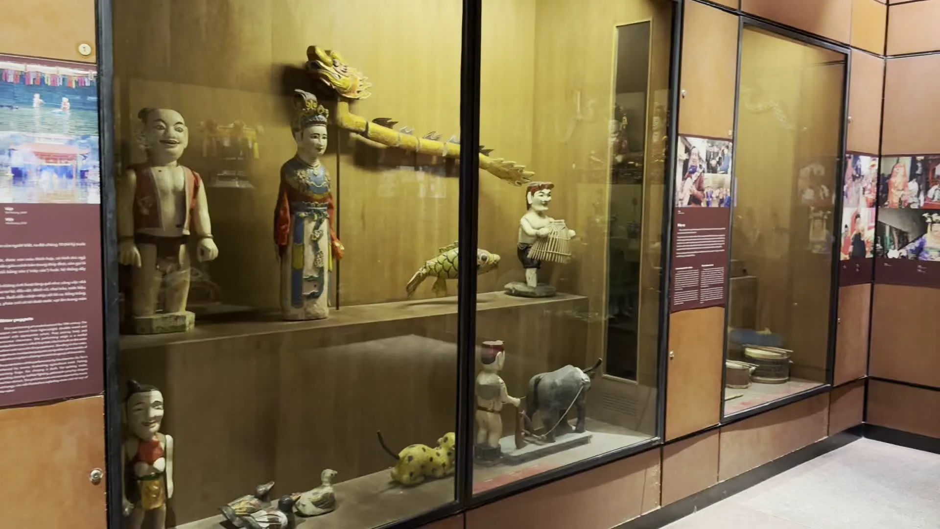 Hình ảnh ghi lại không gian trưng bày bên trong một bảo tàng, có thể là Bảo tàng Dân tộc học Việt Nam. Các hiện vật văn hóa được đặt trong các tủ kính lớn, có khung và nền bằng gỗ màu nâu.。Trong tủ kính đầu tiên, trưng bày các con rối gỗ dùng cho nghệ thuật múa rối nước truyền thống của Việt Nam. Có thể nhận ra các nhân vật quen thuộc như chú Tễu với nụ cười rạng rỡ, một nhân vật hoàng hậu, một con rồng vàng, các con cá, một cậu bé chơi đàn accordion, và một người nông dân đang dắt một con trâu. Ở tầng dưới là các con vịt gỗ.。Ở khu vực trưng bày khác, các tủ kính thể hiện các hình nhân mặc trang phục truyền thống sặc sỡ, có thể liên quan đến các nghi lễ tín ngưỡng dân gian như Hầu đồng. Một số hình nhân mặc áo lụa màu xanh, tím, đỏ, và một hình nhân cầm cây đàn nguyệt. Phía trên có các tượng Phật và các hình trang trí giống con rắn.。Một khu vực khác có tấm bảng ghi rõ tiêu đề "Trò chơi dân gian của trẻ em", cùng với bản dịch tiếng Pháp "Les jeux d'enfants" và tiếng Anh "Children's traditional games". Bên trong tủ kính là nhiều loại mặt nạ, bao gồm cả đầu lân sư tử thường được sử dụng trong dịp Tết Trung Thu, và một mô hình đám rước nhỏ. Tủ kính kế bên trưng bày một bức tranh thờ bằng vải có vẽ các hình và ký tự chữ Hán/Nôm, cùng với các nghiên mực và con dấu.