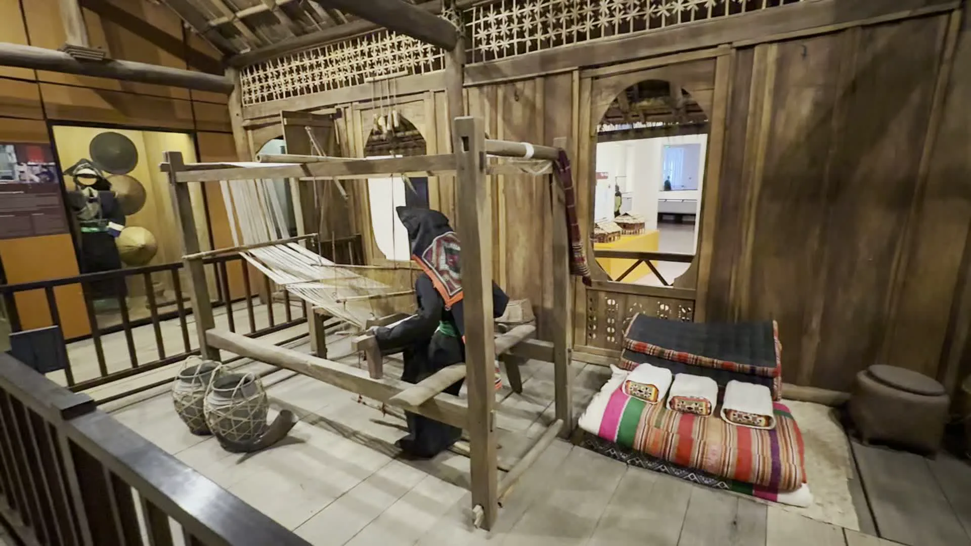 Đây là không gian trưng bày bên trong một bảo tàng, dường như là khu vực tái hiện cuộc sống của một dân tộc thiểu số. Cảnh chính cho thấy một ma-nơ-canh mặc trang phục dân tộc truyền thống đang ngồi bên một khung cửi dệt vải lớn bằng gỗ. Toàn bộ khu vực được dựng lên như một ngôi nhà gỗ, với sàn, tường và các chi tiết trang trí bằng gỗ. Gần đó có hai chiếc gùi đan và một tấm chăn hoặc đệm thổ cẩm nhiều màu sắc được xếp gọn. Trong các hình ảnh khác, có một hành lang bằng gỗ dẫn qua khu trưng bày và các tủ kính chứa các hiện vật như trang phục, nón và các vật dụng sinh hoạt khác. Một người mặc trang phục sáng màu đang đi trong khu vực triển lãm. Không có văn bản hoặc chữ viết nào có thể đọc được.