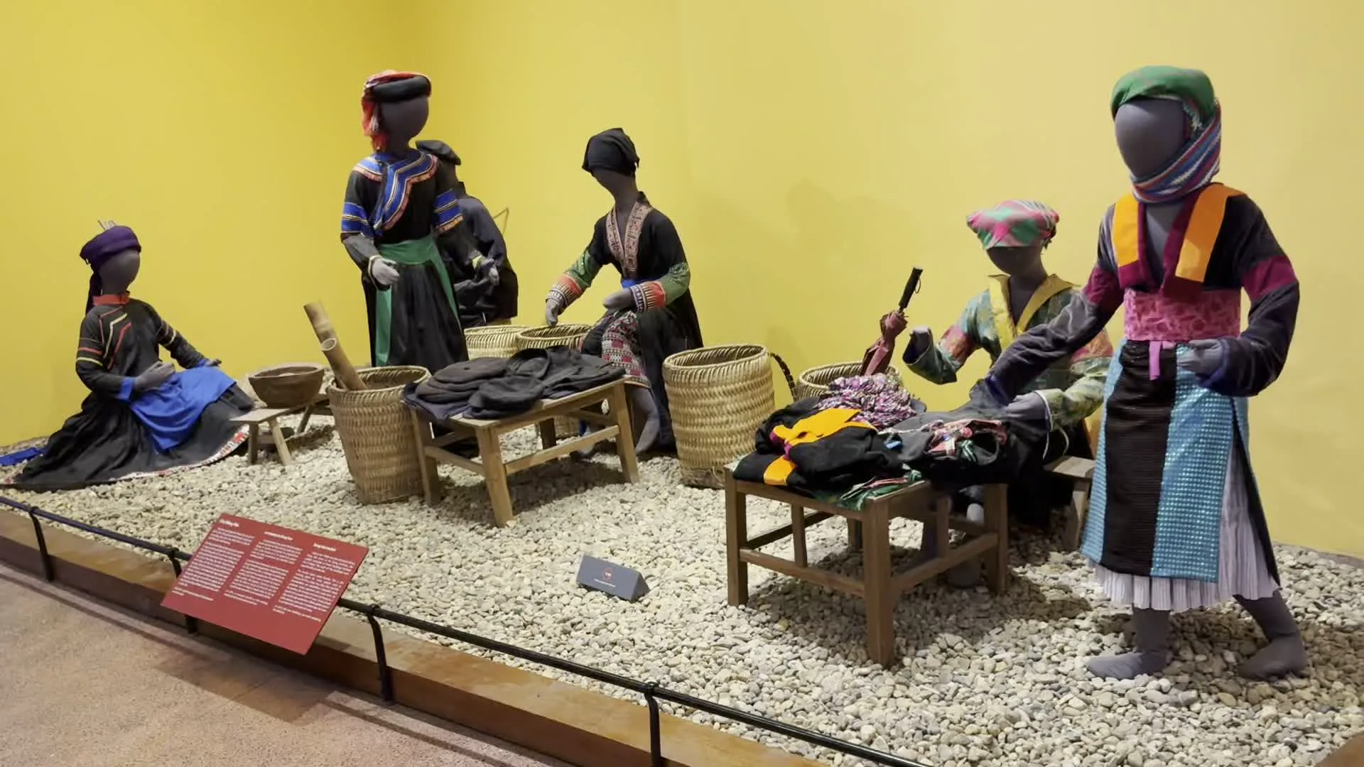 Đây là một khu trưng bày trong bảo tàng, mô phỏng lại một cảnh sinh hoạt của một dân tộc thiểu số. Nhiều ma-nơ-canh được mặc trang phục dân tộc truyền thống, với các họa tiết và màu sắc sặc sỡ, đang quây quần bên những chiếc bàn gỗ thấp. Sàn của khu trưng bày được rải sỏi trắng. Trên bàn có các chồng vải màu sẫm, có thể là vải nhuộm chàm, cùng với những chiếc giỏ, gùi tre đan. Các ma-nơ-canh được sắp đặt trong các tư thế khác nhau: một người đang ngồi, những người khác đang đứng sắp xếp vải hoặc cầm một vật trông giống như dụng cụ để se sợi hoặc dệt vải. Ở góc dưới bên trái có một bảng thông tin màu đỏ, nhưng chữ viết trên đó quá nhỏ và mờ nên không thể đọc rõ nội dung.