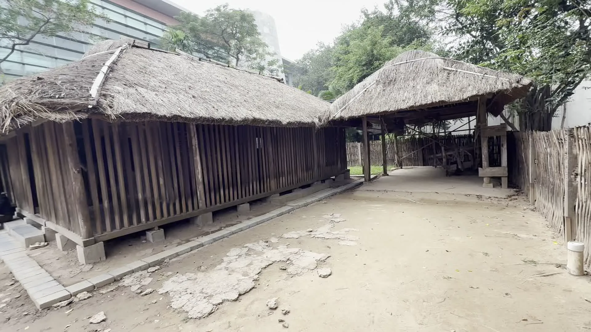 Hình ảnh ghi lại không gian ngoài trời tại một khu trưng bày, có thể là Bảo tàng Dân tộc học Việt Nam, với các công trình kiến trúc truyền thống. Nổi bật là hai ngôi nhà sàn làm bằng gỗ, có mái lợp tranh hoặc lá. Một ngôi nhà dài và một nhà kho hoặc không gian sinh hoạt chung có mái che nối liền. Xung quanh là sân đất, có lối đi lát gạch. Phía sau có thể thấy nhiều cây xanh và một phần của một tòa nhà hiện đại cao tầng. Có một vài khách tham quan trong khu vực: một phụ nữ đang ngồi trên bậc thềm của ngôi nhà sàn, và hai người khác đang đi vào tham quan bên trong một ngôi nhà. Không có văn bản hay chữ viết nào có thể nhìn thấy rõ.