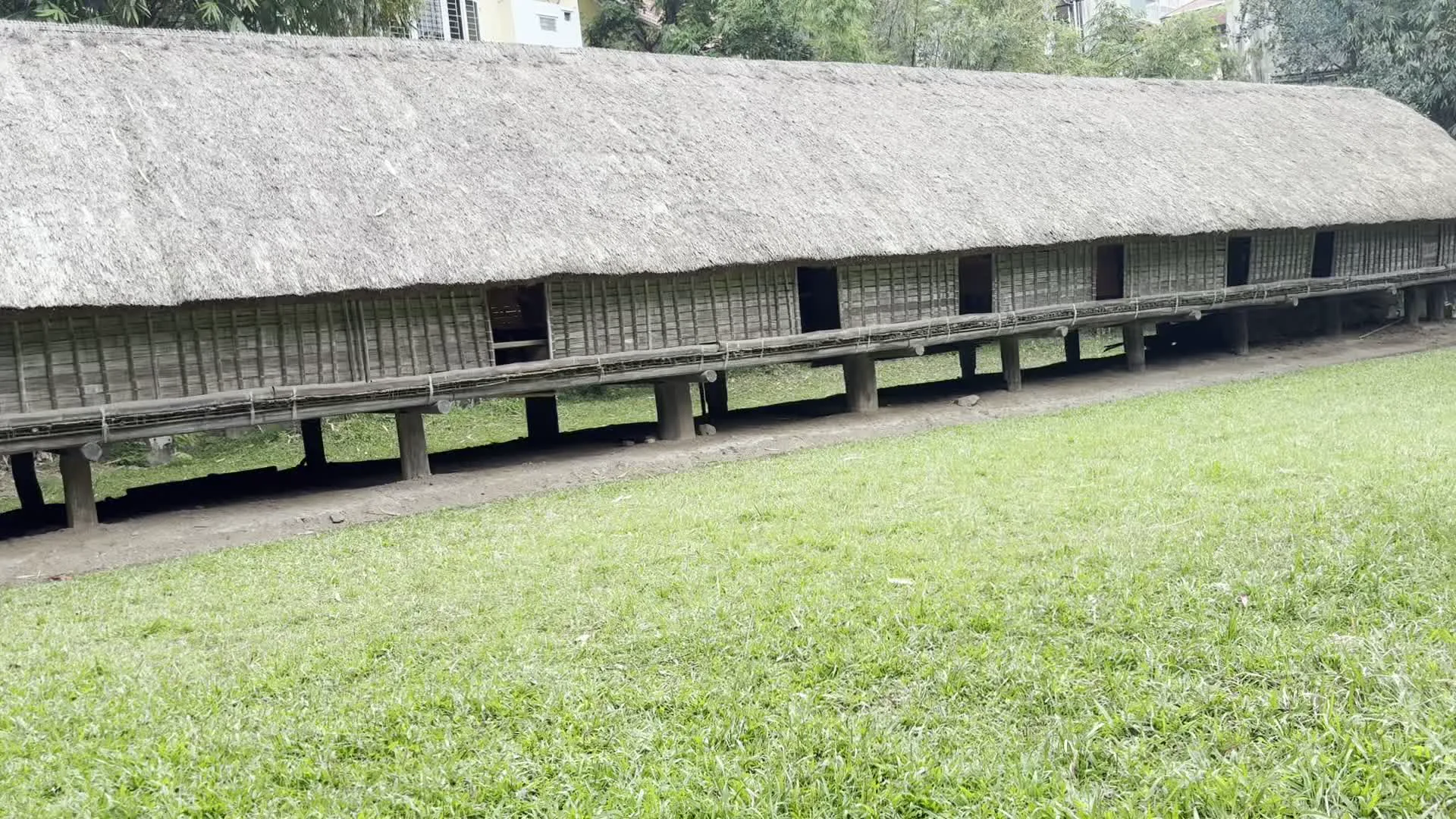 Hình ảnh ghi lại một ngôi nhà sàn truyền thống rất dài, một kiểu kiến trúc đặc trưng của các dân tộc thiểu số ở Việt Nam, có thể là nhà dài của người Ê-đê hoặc M'nông. Ngôi nhà được xây dựng trên nhiều cột gỗ cao và vững chãi, nằm trên một triền dốc cỏ. Mái nhà được lợp bằng vật liệu tự nhiên như cỏ tranh, có độ dày lớn và trải dài suốt toàn bộ công trình. Các bức tường của ngôi nhà được làm bằng vật liệu đan lát, có khả năng là tre hoặc nứa. Có thể thấy nhiều ô cửa sổ hoặc cửa ra vào dọc theo chiều dài của ngôi nhà. Phía trước nhà có một hành lang hẹp chạy dọc theo. Khung cảnh xung quanh là một không gian mở với bãi cỏ xanh và cây cối. Trong một số bức ảnh, có một vài người dường như là du khách đang tham quan, đi lại ở khu vực sân và trên sàn của ngôi nhà. Điều này cho thấy địa điểm có thể là một khu trưng bày ngoài trời trong một bảo tàng dân tộc học hoặc một làng văn hóa du lịch.