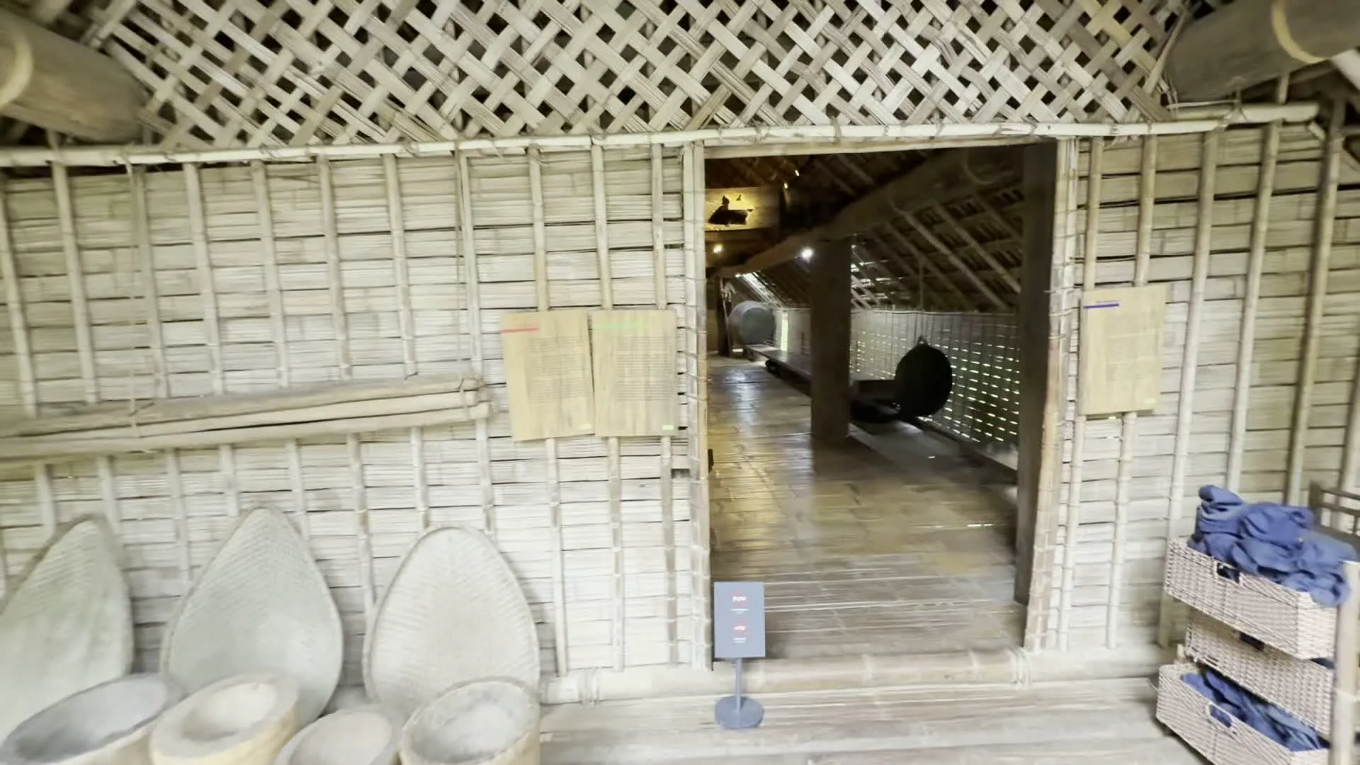 Đây là những hình ảnh ghi lại khung cảnh bên trong và ngoài một khu trưng bày kiến trúc dân tộc, có thể là một bảo tàng như Bảo tàng Dân tộc học Việt Nam. Bên trong là không gian của một ngôi nhà sàn truyền thống, với sàn, tường và mái nhà được làm hoàn toàn từ các vật liệu tự nhiên như gỗ và tre, nứa. Các bức tường được làm bằng phên tre đan. Dọc theo hành lang sàn gỗ, có trưng bày các vật dụng nông nghiệp truyền thống như nia, mẹt (dụng cụ sàng sảy bằng tre) và cối gỗ. Trên tường có treo các bảng thông tin nhưng không thể đọc rõ nội dung. Sự hiện diện của hai bình chữa cháy màu đỏ cho thấy đây là một không gian triển lãm công cộng. Khung cảnh bên ngoài là một khuôn viên rộng lớn với nhiều cây xanh và một lối đi bằng bê tông. Nhiều ngôi nhà sàn khác được nhìn thấy trong khu vực, tạo thành một khu phức hợp trưng bày kiến trúc ngoài trời. Một người đang đi bộ ở phía xa.