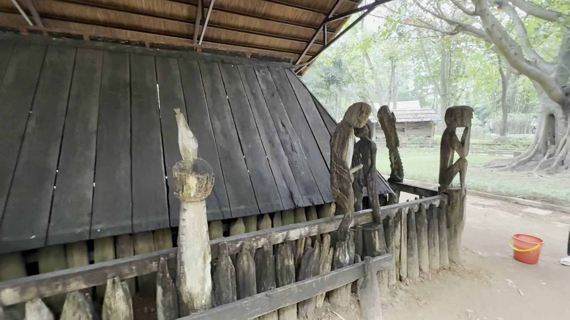 Hình ảnh ghi lại một không gian trưng bày ngoài trời, có thể là trong một bảo tàng hoặc làng văn hóa. Tiêu điểm là một công trình kiến trúc bằng gỗ sẫm màu, giống như một ngôi nhà mồ của một dân tộc thiểu số, có khả năng là của người Gia Rai ở Tây Nguyên. Ngôi nhà mồ được bao quanh bởi một hàng rào gỗ, trên đó có đặt các bức tượng gỗ điêu khắc hình người với nhiều tư thế khác nhau, đây là một đặc trưng trong nghệ thuật tang ma. Toàn bộ khu trưng bày được bảo vệ bởi một mái che lớn có khung bằng kim loại và lợp bằng vật liệu tự nhiên như lá cọ. Có hai du khách, một nam và một nữ, đang đứng xem và tham quan công trình. Xung quanh là không gian nhiều cây xanh. Không có chữ viết, văn bản hay biển hiệu nào trong hình ảnh.