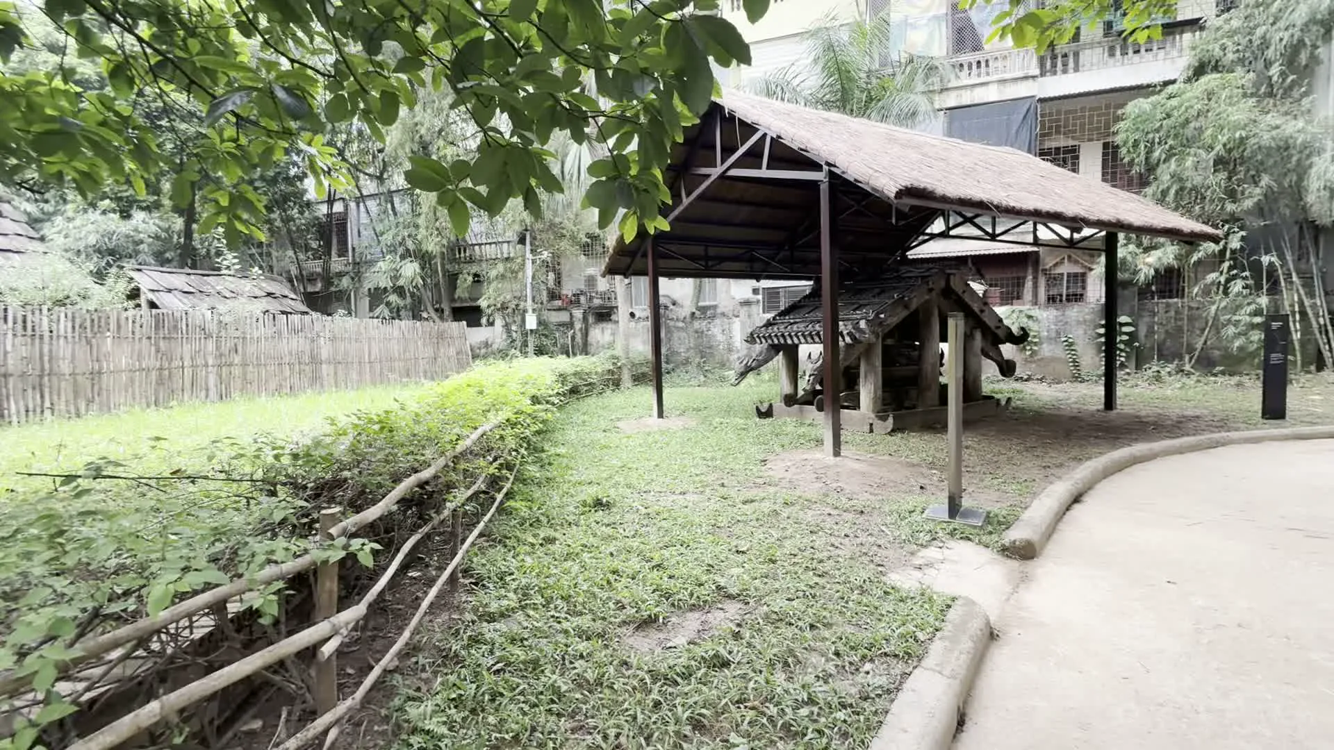 Khung cảnh tại một khu trưng bày ngoài trời, có thể là một bảo tàng dân tộc học, với nhiều cây xanh và bãi cỏ. Có một con đường bê tông uốn lượn dành cho khách tham quan, dọc theo đó là các hàng rào bằng gỗ và ghế đá. Trong khu vực này có trưng bày các công trình kiến trúc truyền thống, bao gồm một ngôi nhà sàn lớn và các ngôi nhà nhỏ lợp mái lá. Nổi bật là một công trình bằng gỗ nhỏ, được chạm khắc tinh xảo, có thể là mô hình nhà mồ, được bảo vệ bởi một mái che riêng lợp lá. Có một vài con gà đang đi lại và kiếm ăn trên bãi cỏ gần công trình này. Phía sau khuôn viên là các tòa nhà cao tầng hiện đại. Có một tấm biển thông tin màu đen đặt trước công trình gỗ nhưng không thể đọc được nội dung.