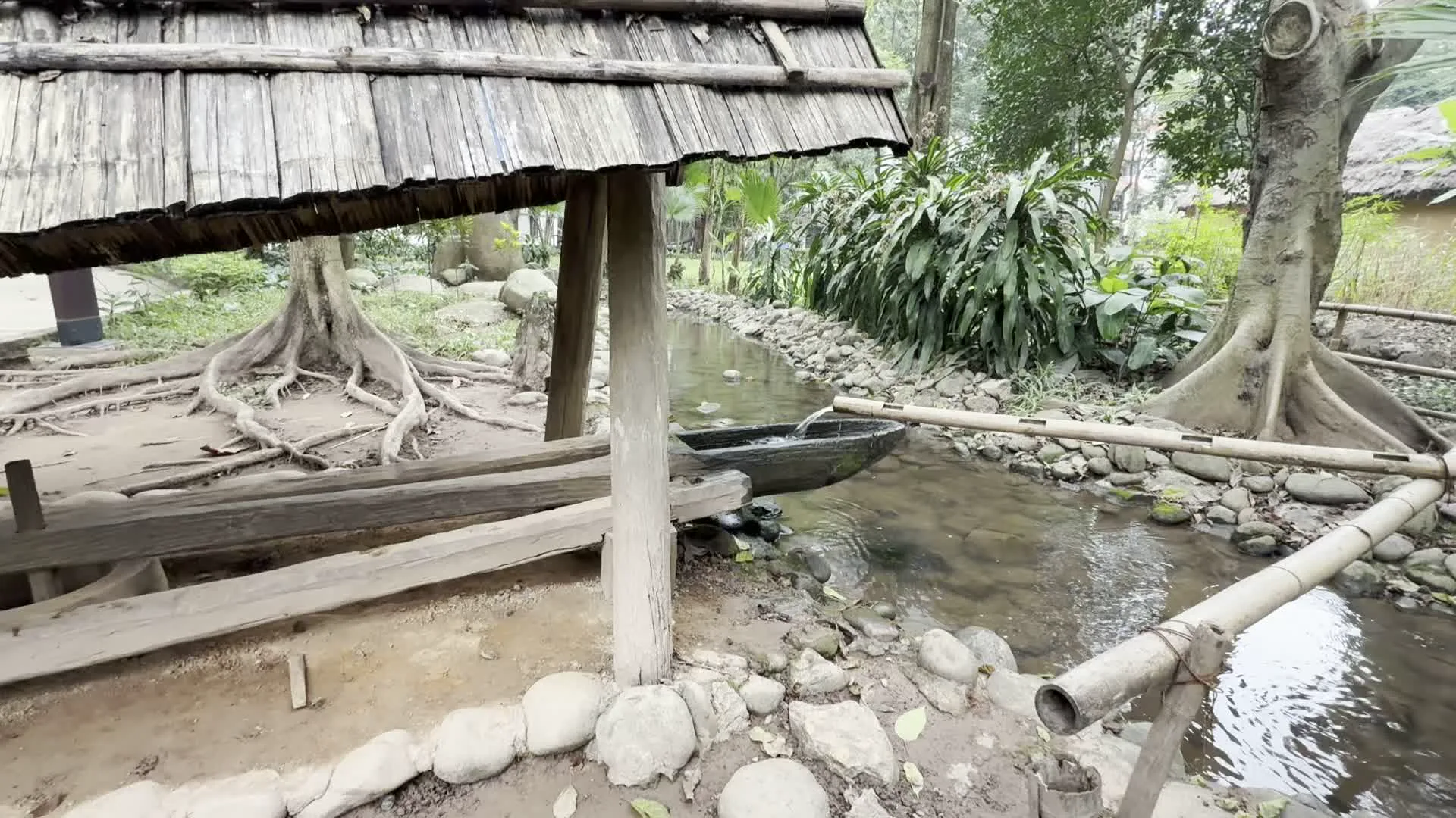 Hình ảnh ghi lại một cảnh quan yên bình tại một con suối nhỏ. Trung tâm của cảnh vật là một hệ thống cối giã gạo truyền thống hoạt động bằng sức nước. Nước được dẫn từ suối qua các ống tre, đổ vào một đầu của cối gỗ có hình dáng giống chiếc thuyền nhỏ. Khi nước đầy, đầu này nặng và chúc xuống, làm đổ nước ra ngoài, khiến đầu kia của cối bật lên rồi giã xuống. Đây là một cơ cấu tự động để giã gạo hoặc các loại nông sản khác. Xung quanh là cây cối xanh tươi, bờ suối được kè bằng đá. Có một mái che đơn sơ làm bằng gỗ và tre được dựng bên cạnh. Ở phía sau, có một cây cầu gỗ bắc qua suối và bóng dáng vài người qua lại.