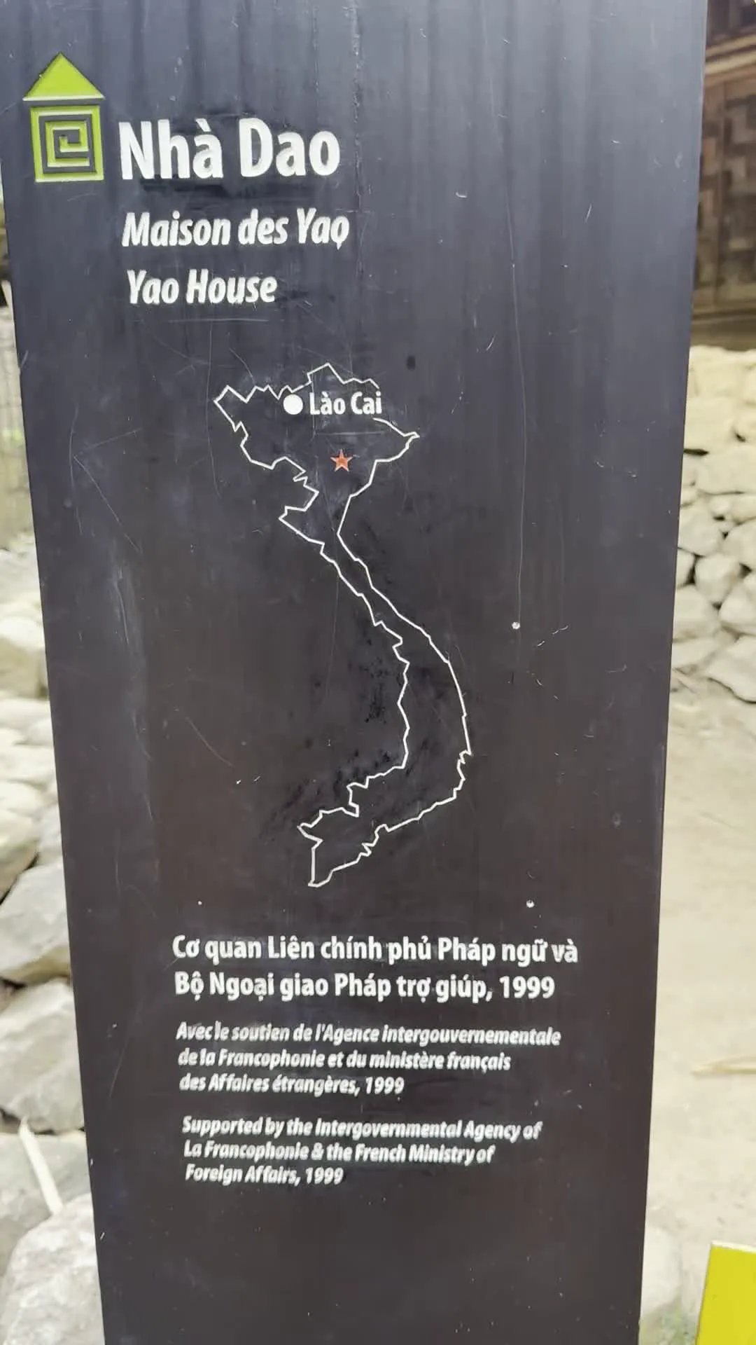 Bức ảnh chụp một tấm biển thông tin màu đen đặt ngoài trời. Trên biển có các nội dung được ghi bằng ba thứ tiếng: Việt, Pháp và Anh.。Nội dung văn bản được ghi lại như sau:。- Tiêu đề: "Nhà Dao", "Maison des Yao", "Yao House".。- Phần giữa: Một bản đồ Việt Nam với tỉnh "Lào Cai" được đánh dấu bằng một ngôi sao.。- Phần dưới: "Cơ quan Liên chính phủ Pháp ngữ và Bộ Ngoại giao Pháp trợ giúp, 1999". Kèm theo đó là bản dịch sang tiếng Pháp ("Avec le soutien de l'Agence intergouvernementale de la Francophonie et du ministère français des Affaires étrangères, 1999") và tiếng Anh ("Supported by the Intergovernmental Agency of La Francophonie & the French Ministry of Foreign Affairs, 1999").。Tấm biển này cung cấp thông tin về một công trình kiến trúc là "Nhà Dao" (Nhà của người dân tộc Dao), tọa lạc tại tỉnh Lào Cai. Công trình này nhận được sự hỗ trợ từ Cơ quan Liên chính phủ Pháp ngữ và Bộ Ngoại giao Pháp vào năm 1999. Phía sau tấm biển có thể thấy một vài tảng đá, có thể là một phần của lối đi hoặc bức tường đá.