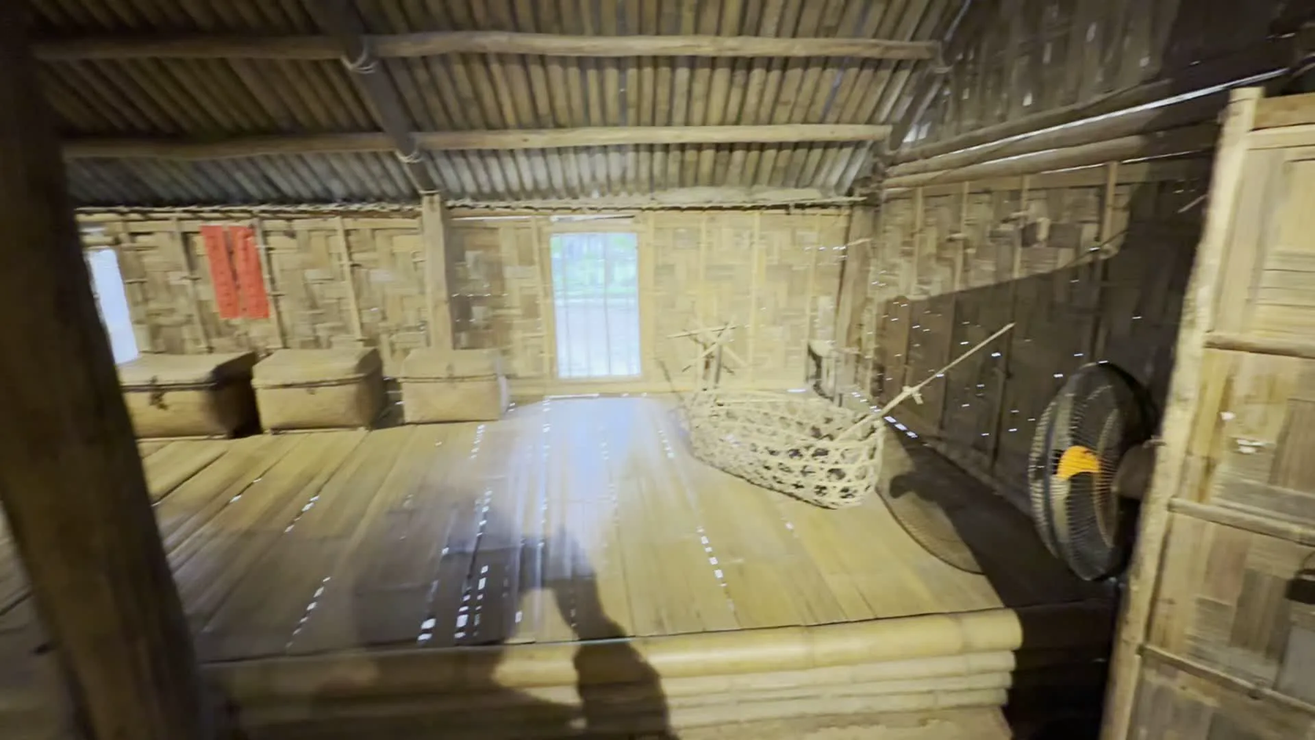 Các hình ảnh cho thấy không gian bên trong một ngôi nhà sàn truyền thống, có khả năng là một khu trưng bày trong bảo tàng hoặc làng văn hóa. Cấu trúc của ngôi nhà chủ yếu làm từ tre và gỗ.。Hình ảnh đầu tiên là một căn phòng có sàn và tường bằng tre. Trong phòng có hai chiếc rương mây tre đan, một chiếc nôi trẻ em cũng bằng tre, và một tấm liễn đỏ có chữ Hán nhưng không thể đọc rõ được treo trên tường. Có một chiếc quạt điện hiện đại ở góc phòng.。Hình ảnh thứ hai là không gian tầng trệt với sàn đất. Trung tâm là một bếp lửa. Phía trên bếp có một cái giàn lớn được treo từ trần nhà, có thể dùng để hong khô hoặc gác đồ. Dọc các bức tường có kệ, chum, vại và một màn hình tivi đang chiếu hình ảnh. Có một biển báo đặt trên giá đỡ nhưng không thể đọc được chữ.。Hình ảnh thứ ba trưng bày một khung dệt cửi thủ công bằng gỗ với các sợi vải trắng đang dệt dở. Gần đó có một tấm bảng chú thích nhưng không đọc được nội dung. Hai du khách đang đứng ở cửa ra vào.