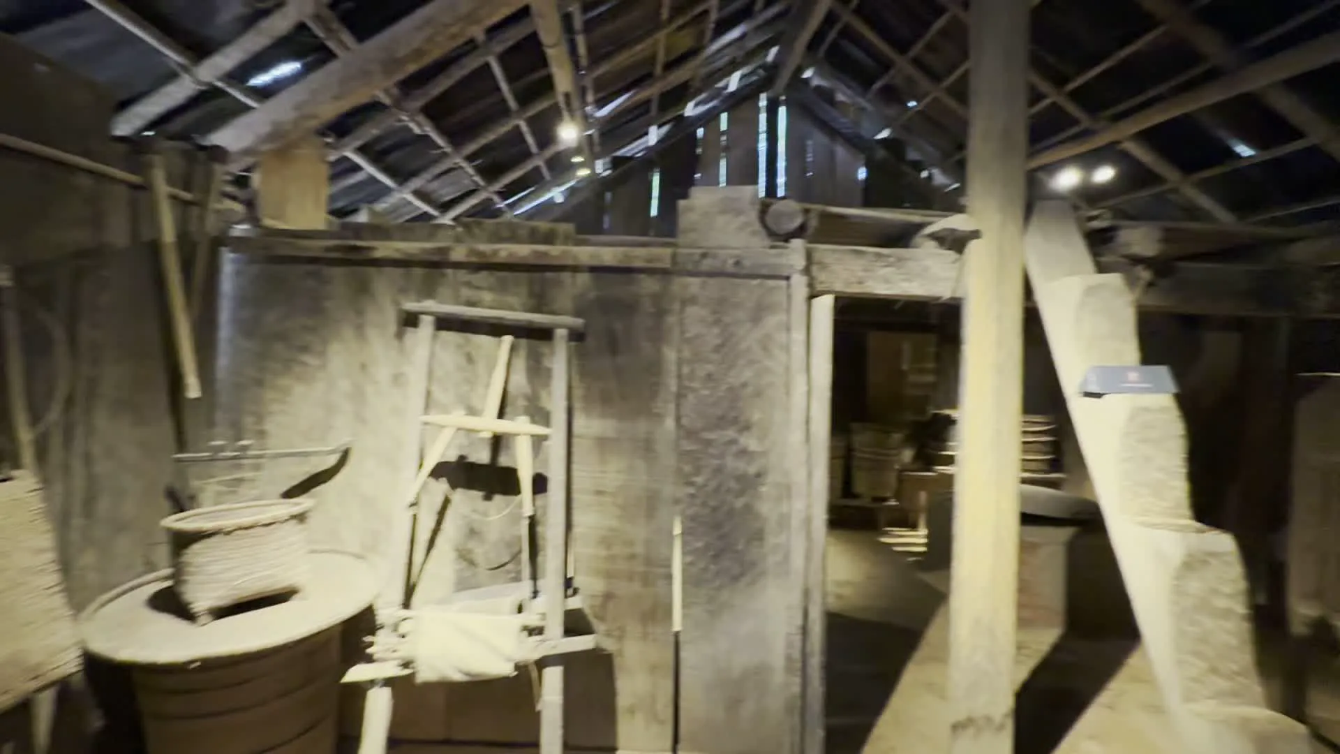 Đây là hình ảnh ghi lại không gian bên trong một ngôi nhà gỗ truyền thống, có thể là một khu trưng bày trong bảo tàng dân tộc học. Toàn bộ kiến trúc như cột, xà, vách ngăn và mái nhà đều được làm bằng gỗ. Sàn nhà dường như là nền đất. Các hình ảnh cho thấy nhiều khu vực khác nhau trong ngôi nhà. Một khu vực có các vật dụng sinh hoạt như một dụng cụ trông giống khung cửi hoặc xa quay sợi và các loại giỏ đan lát. Một gian khác có vẻ là khu vực bếp với các thùng gỗ, chum vại đặt trên kệ và một bếp lò truyền thống. Có cả hình ảnh về một phòng ngủ đơn sơ với giường bằng tấm gỗ, trên tường có treo quần áo và các tấm vải thổ cẩm có hoa văn đặc trưng. Có một vài người dường như là khách tham quan đang đi xem bên trong ngôi nhà.