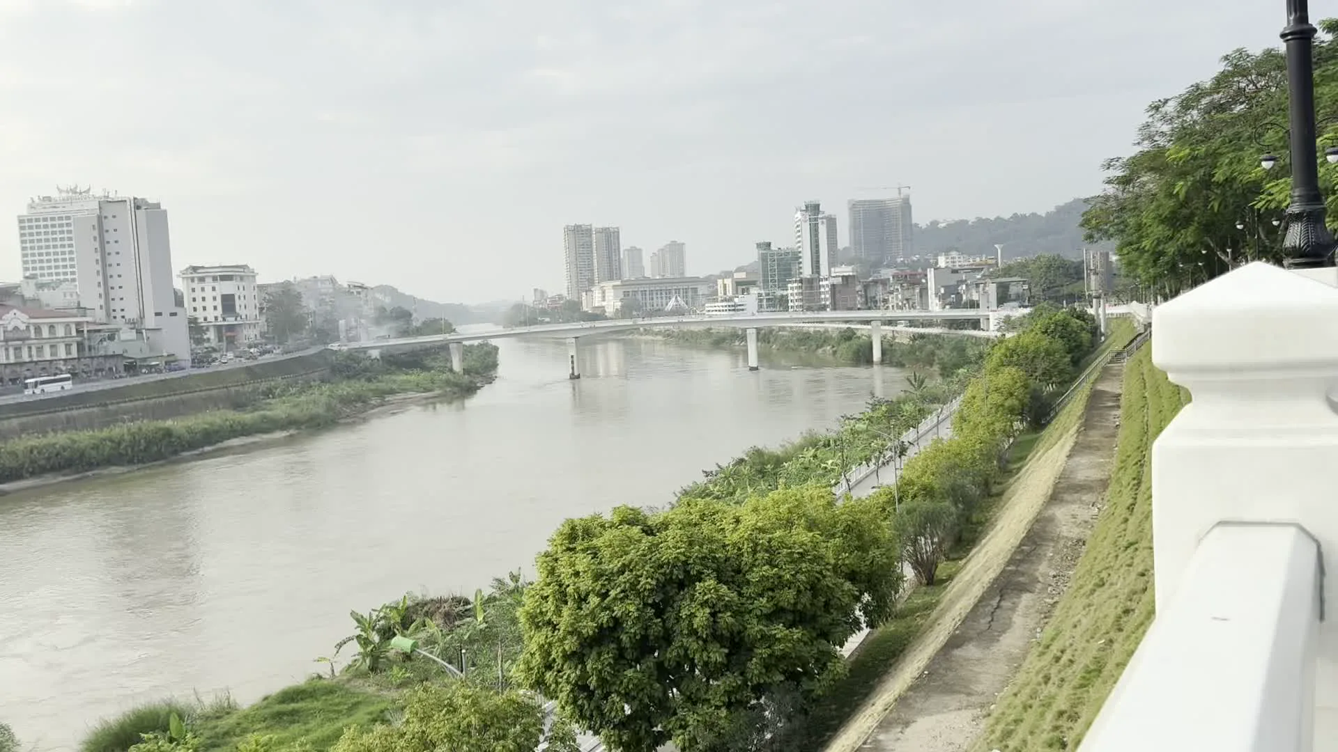 Đây là quang cảnh một thành phố ven sông, có thể là thành phố Lào Cai. Một con sông lớn, nước màu nâu đục, chảy giữa hai khu vực đô thị. Một bên bờ là một thành phố hiện đại với nhiều tòa nhà cao tầng, một số tòa nhà dường như đang được xây dựng. Một cây cầu bê tông dài bắc qua sông. Bờ đối diện là một khu dân cư đông đúc hơn với những ngôi nhà thấp tầng hơn, trong đó có một tòa nhà có kiến trúc giống nhà thờ với cây thánh giá trên đỉnh, và một con đường ven sông có xe cộ qua lại. Có một vài chiếc thuyền nhỏ neo đậu gần bờ. Phía tiền cảnh là một lối đi dạo hoặc công viên ven sông được lát gạch, có lan can màu trắng và các cột đèn đường kiểu cổ. Bờ sông được trồng cây xanh và cỏ. Bầu trời u ám và có nhiều mây.