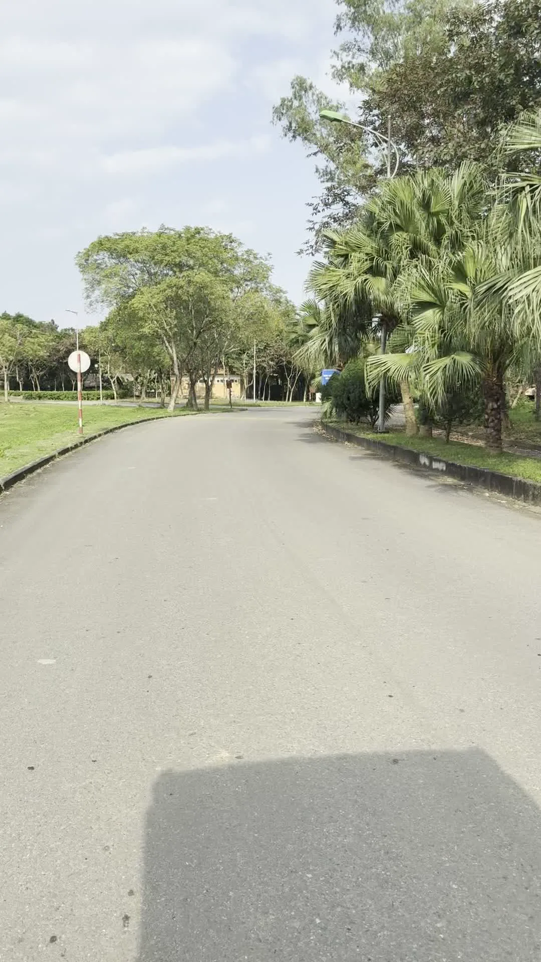 Cảnh quay cho thấy một con đường trải nhựa uốn lượn bên trong một khu vực rộng lớn có nhiều cây xanh. Hai bên đường là bãi cỏ và các loại cây đa dạng như cây cọ và cây thân gỗ. Một chiếc xe điện du lịch màu trắng đang đỗ ở lề đường. Trên kính chắn gió của xe có dòng chữ màu xanh lá cây 'LÀNG VĂN HÓA - DU LỊCH CÁC DÂN TỘC VIỆT NAM'. Cạnh đó có một cột cờ treo lá cờ đỏ sao vàng của Việt Nam. Ngoài ra còn có một biển báo giao thông hình tròn viền đỏ và hệ thống đèn đường dọc theo con đường. Khung cảnh vắng vẻ, cho thấy một không gian yên tĩnh trong một ngày trời trong.