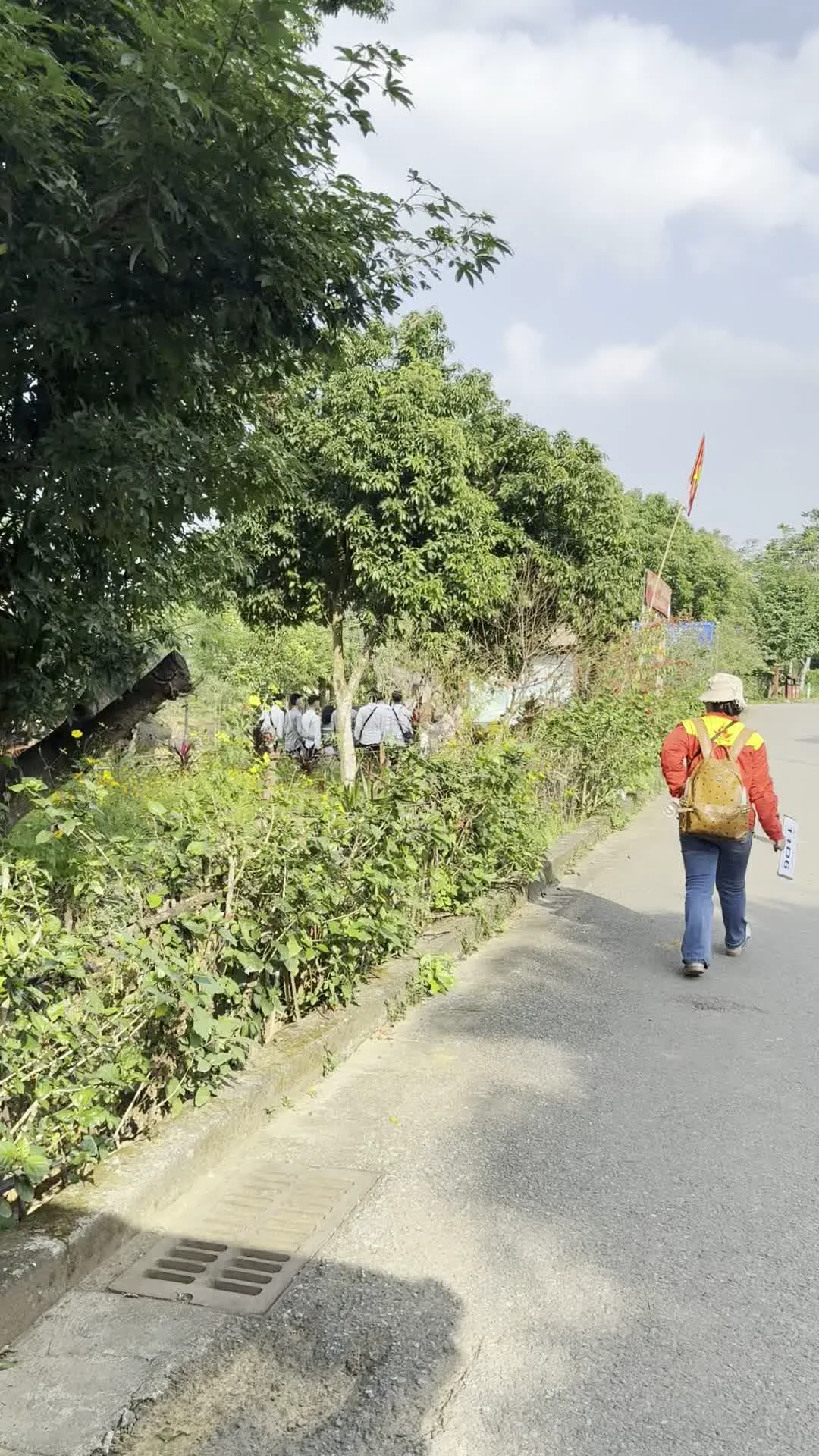 Bản ghi văn bản: Một người trong đoàn đang cầm một tấm biển có ghi chữ 'TRẠM 5'.。Mô tả cảnh vật và hoạt động: Khung cảnh diễn ra ngoài trời, ban ngày, tại một khu vực có vẻ là một khu di tích lịch sử hoặc một công viên. Một nhóm người đang đi bộ tham quan dọc theo một con đường nhựa. Bên đường là hàng rào cây xanh và cây cối um tùm. Một người phụ nữ đeo ba lô, mặc áo khoác đỏ, đang đi bộ và cầm tấm biển ghi 'TRẠM 5'. Phía xa hơn có một người đang cầm lá cờ Việt Nam. Các hình ảnh khác cho thấy một con đường chạy dọc theo một bãi cỏ có trồng hàng cây cọ, và một hồ nước lớn, phẳng lặng. Phía bên kia hồ là một tòa nhà lớn với mái ngói đỏ, được bao quanh bởi cây xanh, và hình ảnh của nó được phản chiếu trên mặt nước.