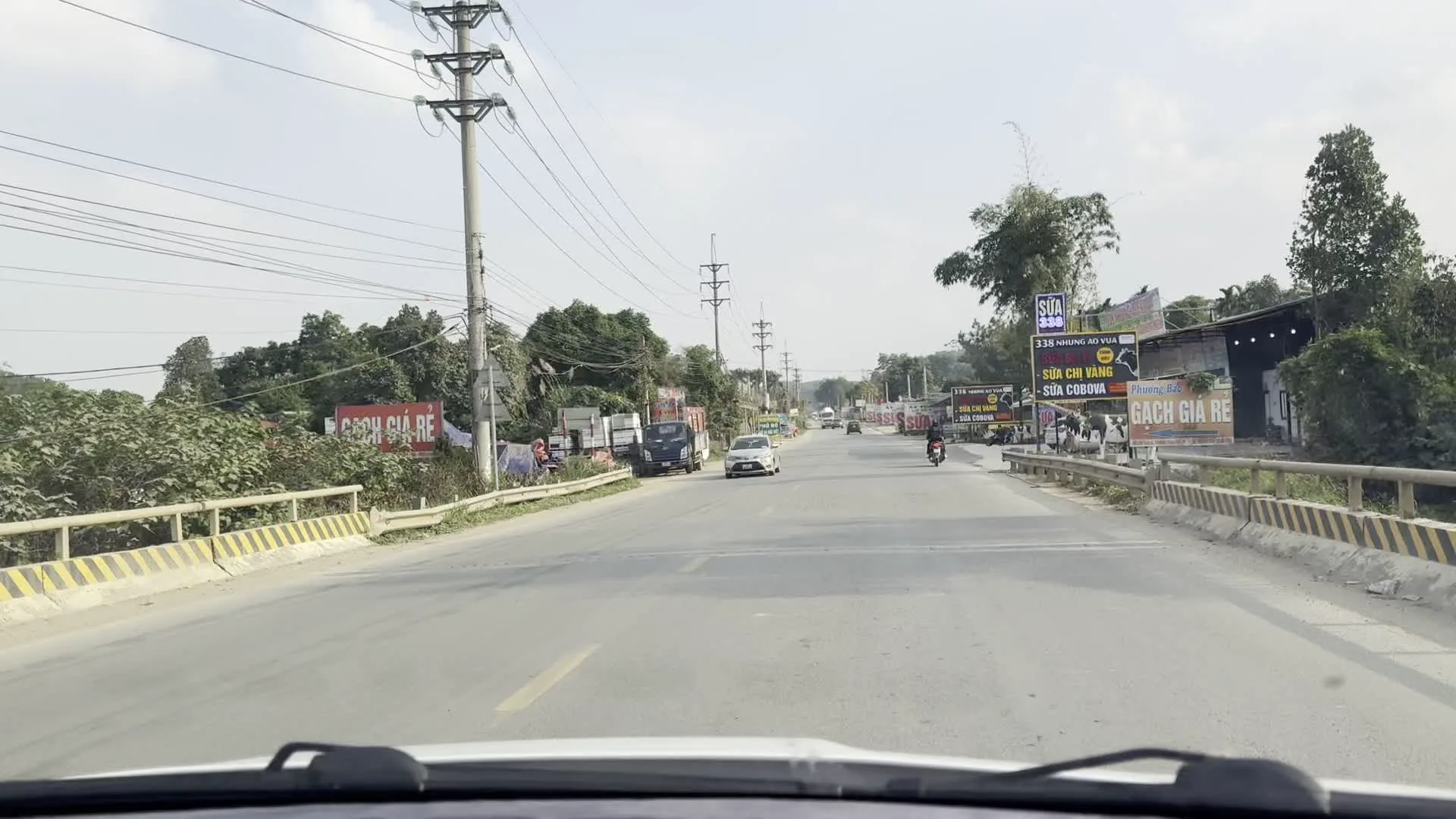 Quang cảnh được ghi lại từ góc nhìn bên trong một chiếc ô tô đang di chuyển trên một con đường quốc lộ hoặc tỉnh lộ ở Việt Nam. Con đường được trải nhựa khá rộng, hai bên là nhà dân và các cửa hàng kinh doanh. Dọc đường có nhiều cột điện, dây điện chằng chịt và cây cối xanh tươi.。Hoạt động chính là giao thông với nhiều loại xe như ô tô, xe máy, xe tải và xe buýt đang lưu thông. Các cửa hàng hai bên đường đang hoạt động, được thể hiện qua hàng loạt biển hiệu quảng cáo.。Nhiều biển hiệu và biểu ngữ được treo lên, các văn bản có thể nhận dạng bao gồm:。- Biển quảng cáo màu đỏ: "GẠCH GIÁ RẺ".。- Biển quảng cáo sản phẩm sữa: "SỮA BA VÌ", "Sữa chua", "CÔNG TY CỔ PHẦN SỮA TƯƠI BA VÌ", "ĐẶC SẢN SỮA".。- Biển của một đại lý: "ĐẠI LÝ ÁT THẢO Kim An" quảng cáo "SỮA + BÒ + DÊ", "100% SỮA TƯƠI".。- Biển quảng cáo khác: "SIM 3G/4G", "336 NHƯỢNG HỒ VÀNG...".。- Một biển hiệu có số điện thoại: "0981.700 - 098.943.9999".。Một lá cờ Việt Nam (cờ đỏ sao vàng) được treo trên một biển hiệu lớn. Hình ảnh cũng cho thấy một cây cầu có lan can bê tông sơn sọc vàng.