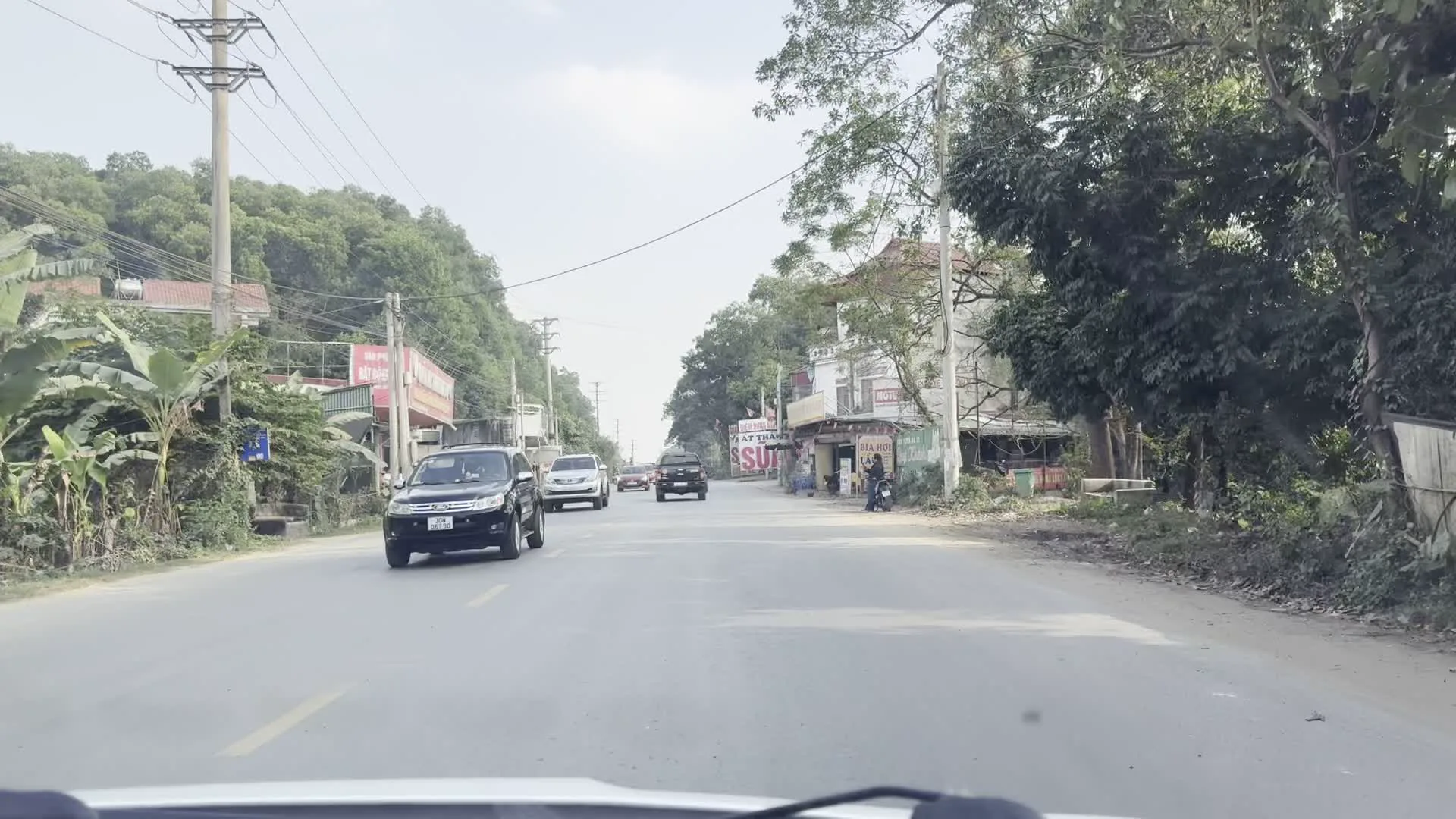 Khung cảnh được ghi lại từ góc nhìn của một chiếc xe ô tô đang di chuyển trên một con đường ở vùng nông thôn Việt Nam. Con đường được trải nhựa và có nhiều xe ô tô khác đang lưu thông. Hai bên đường là cây cối xanh tươi, những ngọn đồi và các tòa nhà. Phía bên phải đường là một khu vực dường như là trạm dừng nghỉ, với nhiều biển hiệu quảng cáo. Các biển hiệu có thể đọc được bao gồm: "TRẠM DỪNG NGHỈ ĐẶT THẢO", "SỮA BÒ DÊ", "CƠM XE TẢI", "ĐIỂM BÁN ĐẶC SẢN", "GIÒ BÒ", và "SỮA CHUA". Điều này cho thấy đây là nơi bán các đặc sản địa phương như sữa tươi, sữa chua, giò bò và phục vụ các bữa ăn cho khách đi đường, đặc biệt là tài xế xe tải. Có một vài người và xe máy đang ở khu vực các cửa hàng ven đường.