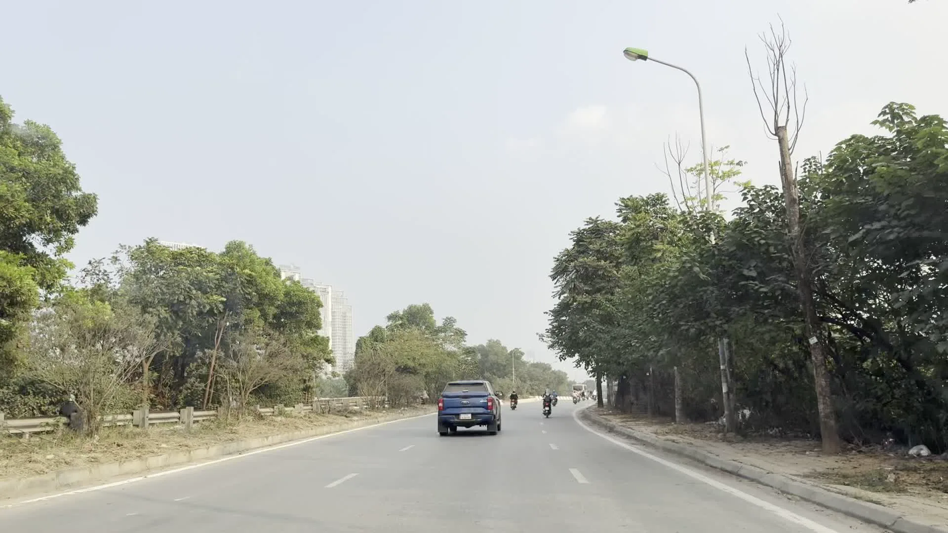 Khung cảnh được ghi lại từ bên trong một chiếc ô tô đang di chuyển trên một con đường lớn, nhiều làn xe tại Việt Nam. Trên đường có nhiều phương tiện giao thông khác nhau như ô tô và xe máy. Phía trước có một chiếc xe bán tải màu xanh lam. Hai bên đường được trồng nhiều cây xanh và có lan can bảo vệ. Ở phía xa, có thể nhìn thấy một tòa nhà cao tầng, cho thấy đây là khu vực đô thị hoặc ven đô. Những người đi xe máy đội mũ bảo hiểm, một số người đeo khẩu trang. Dọc đường có các cột đèn chiếu sáng. Một hình ảnh cho thấy có biển báo cấm đi ngược chiều (hình tròn màu đỏ, vạch ngang màu trắng). Có một hàng rào tôn màu xanh lá cây bên đường với các hình vẽ hoặc chữ viết phun sơn nhưng không thể đọc rõ. Một hình ảnh khác chụp cận cảnh nội thất ô tô, cho thấy cửa gió điều hòa trên bảng điều khiển và một tấm thảm phủ taplo.