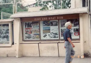 Trên một bảng thông tin bằng gỗ phía trên tủ trưng bày có dòng chữ lớn 'HINH ANH NHAT BAN'. Đây là một khu vực trưng bày ảnh ngoài trời trên vỉa hè ở Hà Nội, chụp vào năm 1992. Tủ trưng bày được xây dựng kiên cố, có mái che và mặt kính, bên trong trưng bày nhiều bức ảnh màu và đen trắng về đất nước, con người và công nghệ Nhật Bản. Có thể thấy các hình ảnh về tàu cao tốc, núi Phú Sĩ, ô tô, và các hoạt động của người dân. Một người đàn ông lớn tuổi, tóc bạc, mặc áo sơ mi cộc tay màu xanh và quần dài, đang đi bộ trên vỉa hè ngang qua khu trưng bày. Phía sau là hàng rào lưới và cây cối.。（日本語訳）。ショーケースの上にある木製の案内板には、「HINH ANH NHAT BAN」という大きな文字が書かれている。これは1992年にハノイの歩道で撮影された屋外写真展示エリアである。ショーケースは頑丈に作られており、屋根とガラス面があり、中には日本の国土、人々、技術に関する多くのカラー写真や白黒写真が展示されている。白髪で、青い半袖シャツと長ズボンを履いた年配の男性が、展示エリアの横の歩道を歩いている。後ろには金網のフェンスと木々がある。