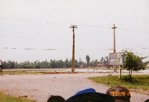 Bức ảnh chụp một lối vào có cổng của một khu nhà hoặc cơ quan, phản ánh khung cảnh đặc trưng của Việt Nam vào năm 1992.。Loại địa điểm: Đây là cổng vào của một khu vực có tường rào, có thể là một cơ quan, nhà máy, hoặc một khu tập thể. Sân được lát gạch block (gạch con sâu), nhưng nhiều chỗ đã bị bong tróc, hư hỏng và không bằng phẳng.。Các vật thể trong ảnh:。- Một chiếc cổng sắt lớn, sơn màu xanh đậm, đang ở trạng thái đóng.。- Hai cột trụ lớn, xây bằng gạch và sơn màu vàng, đỡ một dầm bê tông có mái che nhỏ kiểu cách điệu ở phía trên.。- Phía sau cổng, có một chiếc ô tô con màu trắng đang đỗ.。- Một người đàn ông mặc áo phông sáng màu và quần jean xanh đang đứng cạnh chiếc xe.。- Ở hậu cảnh là một dãy nhà cấp bốn, tường trắng, lợp mái tôn hoặc fibro xi măng đã cũ và có vài chỗ vá.。- Bên phải có một cây xanh lớn, rậm rạp.。- Một hàng rào sắt chạy dọc theo khuôn viên.。Hoạt động: Khung cảnh tĩnh lặng. Một người đang đứng bên trong khuôn viên, gần chiếc ô tô. Cổng đang đóng, cho thấy sự ra vào được kiểm soát.。Chữ viết: Không có chữ viết nào có thể đọc được trong ảnh.。（日本語訳）。写真は、1992年のベトナムの典型的な風景を反映した、ある住宅地または機関の門のある入り口を写している。場所の種類：これは壁で囲まれたエリアの入り口の門で、機関、工場、または集合住宅である可能性がある。中庭はインターロッキングブロック（芋虫レンガ）で舗装されているが、多くの場所が剥がれ、損傷し、平らではない。写真の中の物体：。- 濃い緑色に塗られた大きな鉄の門が、閉まった状態である。- レンガで造られ黄色く塗られた2本の大きな柱が、上部に様式化された小さな屋根が付いたコンクリートの梁を支えている。- 門の後ろに、白い乗用車が1台駐車している。- 明るい色のTシャツと青いジーンズを履いた男性が、車の隣に立っている。- 背景には、白い壁の平屋の家々が並んでおり、古いトタンまたはファイバーセメントで屋根が葺かれ、数カ所補修されている。- 右側には、大きな緑の木が鬱蒼と茂っている。- 鉄の柵が敷地に沿って続いている。活動：静かな光景。一人の人物が敷地内で、車の近くに立っている。門は閉まっており、出入りが管理されていることを示している。文字：写真の中には読み取れる文字はない。