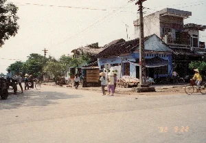 Bức ảnh ghi lại quang cảnh đường phố ở một vùng quê hoặc thị trấn của Việt Nam vào ngày 22 tháng 9 năm 1992. Con đường được trải nhựa, hai bên là những ngôi nhà với kiến trúc đan xen giữa truyền thống và hiện đại. Nổi bật là một ngôi nhà cấp bốn sơn màu xanh lam với mái hiên, bên cạnh và phía sau là những ngôi nhà lợp ngói và một ngôi nhà tầng bê tông. Hệ thống cột điện và dây điện chằng chịt phía trên. Hoạt động của con người diễn ra khá tấp nập: một nhóm phụ nữ đội nón lá đang đi bộ trên đường, một người phụ nữ khác cũng đội nón lá đang đi xe đạp. Ở bên trái, có một chiếc xe kéo bằng gỗ (có thể là xe bò) và một vài người đang tụ tập xung quanh. Ven đường, có một sạp hàng nhỏ bày bán sản phẩm, cho thấy không khí sinh hoạt và buôn bán của địa phương. Có một biển báo giao thông hình tròn nhưng không thể nhận dạng được biểu tượng. Trên ban công của ngôi nhà tầng có một tấm biển chữ vàng nhưng không thể đọc rõ nội dung. Dấu ngày tháng trên ảnh ghi rõ '92 9 22.。（日本語訳）。この写真は1992年9月22日のベトナムの田舎または町における街の風景を記録したものです。道路は舗装されており、両側には伝統と現代が混在した建築様式の家々が並んでいます。目立っているのは、軒先のある青い平屋の家で、その隣と後ろには瓦葺きの家々とコンクリート造りの階建ての家があります。頭上には電柱と電線が張り巡らされています。人々の活動はかなり活発です。ノンラー（菅笠）をかぶった女性の一団が道を歩いており、同じくノンラーをかぶった別の女性が自転車に乗っています。左側には、木製の荷車（牛車かもしれません）があり、その周りに数人が集まっています。道端には商品を並べた小さな屋台があり、地域の生活や商売の様子がうかがえます。円形の交通標識がありますが、その記号は識別できません。階建ての家のバルコニーには黄色い文字の看板がありますが、内容ははっきりと読み取れません。写真の日付スタンプには「'92 9 22」と明記されています。