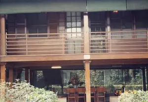 Hình ảnh chụp một ngôi nhà sàn hai tầng làm bằng gỗ, một kiểu kiến trúc đặc trưng ở Việt Nam. Tầng trên có ban công với lan can gỗ, cửa sổ kính khung gỗ và cửa chớp. Phía trên có mái hiên hoặc rèm che màu xanh lá. Tầng trệt là một không gian mở, được chống đỡ bởi các cột gỗ. Dưới gầm sàn, có một bộ bàn ghế gỗ dài được sắp xếp gọn gàng, trông giống như một phòng họp hoặc nơi tiếp khách ngoài trời. Trên bàn có một bình hoa lớn được cắm công phu. Phía sau là một khu vườn với nhiều cây xanh. Có một tấm biển nhỏ màu trắng gắn trên một cây cột nhưng không thể đọc được nội dung. Bối cảnh và kiến trúc của ngôi nhà gợi liên tưởng mạnh mẽ đến khu di tích Nhà sàn Bác Hồ tại Hà Nội.。（日本語訳）。木造二階建ての高床式家屋を写した写真で、ベトナムの典型的な建築様式です。上階には木製の手すりが付いたバルコニー、木枠のガラス窓、そして鎧戸があります。その上には緑色の軒、または日よけのカーテンがあります。1階はオープンスペースで、木製の柱で支えられています。床下には、長い木製のテーブルと椅子のセットがきちんと配置されており、会議室や屋外の応接スペースのように見えます。テーブルの上には、精巧に生けられた大きな花瓶があります。後ろには緑豊かな庭があります。柱には小さな白い看板が取り付けられていますが、内容は読み取れません。この家の背景と建築は、ハノイにあるホーチミン主席の高床式の家の史跡を強く連想させます。