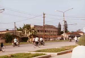 Trên mặt tiền của tòa nhà lớn ở phía sau có dòng chữ: 'NHÀ MÁY CHẾ TẠO MÁY CÔNG CỤ SỐ 1'. Trên một bức tường ở bên trái cũng có chữ viết nhưng khó đọc, chỉ nhận ra được chữ 'CHUYÊN...'.。Bối cảnh là một con đường phố ở Hà Nội vào năm 1992. Con đường được trải nhựa, có dải phân cách trồng cỏ ở giữa. Dọc hai bên đường là các tòa nhà, trong đó nổi bật là một nhà máy lớn, nhiều tầng. Phía trước nhà máy là các dãy nhà cấp bốn lợp mái tôn đã gỉ sét.。Hoạt động chính được ghi lại là cảnh giao thông trên đường. Nhiều người đang di chuyển bằng xe đạp và xe máy. Các phương tiện này là phương tiện đi lại phổ biến của người dân thời kỳ đó. Một người ở phía trước, góc phải, đang đội một chiếc mũ cối màu xanh lá cây.。Các vật thể có trong ảnh bao gồm: tòa nhà 'Nhà máy Chế tạo Máy công cụ số 1', xe đạp, xe máy (kiểu dáng cũ như Honda Cub), một chiếc ô tô kiểu xe van đỗ ven đường, cột điện, cột đèn đường và hệ thống dây điện, dây cáp chằng chịt.。（日本語訳）。後方にある大きな建物の正面には、「第一工作機械製造工場」という文字がある。左側の壁にも文字が書かれているが、読みづらく、「CHUYÊN...」という文字しか認識できない。舞台は1992年のハノイのある通りである。道路はアスファルトで舗装されており、中央には草が植えられた分離帯がある。道路の両脇には建物が並んでおり、その中でもひときわ目立つのは、大きな多層階の工場である。工場の前には、錆びたトタン屋根の平屋建ての家々が並んでいる。記録されている主な活動は、道路上の交通風景である。多くの人々が自転車やバイクで移動している。これらの乗り物は、当時の人々の一般的な交通手段であった。前景の右隅にいる一人の人物が、緑色のヘルメットをかぶっている。写真に含まれる物体には、「第一工作機械製造工場」の建物、自転車、バイク（ホンダ・カブのような旧式）、道端に駐車されたバンタイプの車、電柱、街灯、そして複雑に絡み合った電線やケーブルのシステムが含まれる。