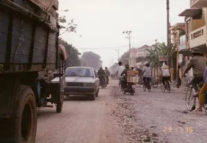 Bức ảnh ghi lại khung cảnh giao thông trên một con đường đất, gồ ghề và đang trong tình trạng xuống cấp ở một khu vực có thể là ngoại ô Hà Nội vào năm 1992. Bên trái là một phần của chiếc xe tải lớn, cũ kỹ. Trên đường có nhiều người đang di chuyển bằng xe đạp, một số người đội nón lá, một người đàn ông đội mũ cối. Một chiếc ô tô con màu sáng đang đi tới. Ngoài ra còn có một chiếc xe máy đang chở hai người cùng với các thùng hàng carton được buộc ở phía sau. Dọc bên phải đường là các ngôi nhà, cột điện và cây cối. Có một biển hiệu gắn trên cột điện ghi chữ 'BƯU ĐIỆN TRUNG ƯƠNG'. Một biển hiệu khác màu xanh trắng gắn trên một ngôi nhà nhưng chữ viết không thể đọc rõ.。（日本語訳）。この写真は、1992年、ハノイ郊外と思われる地域にある、未舗装ででこぼこし、劣化が進んでいる道路の交通風景を記録したものです。道路上では多くの人々が自転車で移動しており、何人かはノンラー（菅笠）をかぶり、一人の男性は軍用ヘルメットをかぶっています。明るい色の乗用車が1台、こちらに向かってきています。その他に、2人乗りで、後ろに段ボール箱をいくつも縛り付けているバイクが1台あります。道路の右側沿いには、家々、電柱、そして木々が並んでいます。電柱には「BƯU ĐIỆN TRUNG ƯƠNG」（中央郵便局）と書かれた看板が取り付けられています。別の青と白の看板が家に取り付けられていますが、文字ははっきりと読み取れません。