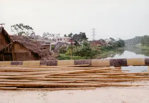 Cảnh quan một khu vực ven sông, có thể được chụp từ trên một cây cầu hoặc một con đường ven đê. Ở phía trước, một đống lớn các cây tre, trúc được chất đống trên mặt đất, có thể là vật liệu xây dựng. Ngay sau đó là một lan can bê tông được sơn sọc vàng và đen. Bên kia lan can là một con sông hoặc kênh rạch. Phía bờ đối diện là một khu dân cư với nhiều loại nhà cửa khác nhau. Nổi bật ở bên trái là một ngôi nhà tranh, vách đất hoặc tre, có mái lợp bằng lá, một kiểu nhà truyền thống. Phía sau và xung quanh là những ngôi nhà xây kiên cố hơn, một số là nhà tầng. Cây cối xanh tươi mọc dọc theo bờ sông. Một cột điện cao thế cũng có thể được nhìn thấy ở phía xa. Không có hoạt động nào của con người được ghi lại rõ ràng. Ở góc dưới bên phải của bức ảnh có một dấu ngày tháng mờ, có thể đọc là '29 9 '92'.。（日本語訳）。川沿いの地域の風景で、橋の上か堤防沿いの道から撮影されたものと思われる。手前には、大量の竹が地面に積まれており、建築資材かもしれない。そのすぐ後ろには、黄色と黒の縞模様に塗られたコンクリートの欄干がある。欄干の向こう側は川か運河である。対岸は様々な種類の家々が建ち並ぶ住宅地である。左側で目立っているのは、土壁か竹壁で、屋根が葉で葺かれた茅葺きの家で、伝統的な様式の家である。その後ろや周りには、より頑丈に建てられた家々があり、いくつかは階建てである。青々とした木々が川岸に沿って生えている。遠くには高圧電線の鉄塔も見ることができる。人間の活動ははっきりと記録されていない。写真の右下隅には、「'92 9 29」と読めるぼやけた日付の印がある。