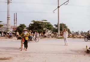 Bức ảnh ghi lại một cảnh đường phố ở Việt Nam, có thể là khu vực ngoại ô Hà Nội, vào khoảng năm 1992. Dưới góc phải bức ảnh có dấu thời gian kỹ thuật số màu cam ghi '29 17:47'.。Đây là một con đường hoặc ngã tư khá đông đúc với mặt đường bằng đất, chưa được trải nhựa hoàn chỉnh. Một người cảnh sát giao thông trong bộ đồng phục trắng đang đứng giữa đường, dường như đang làm nhiệm vụ điều tiết giao thông. Dòng người và phương tiện qua lại tấp nập, chủ yếu là xe đạp và xe máy, loại phương tiện phổ biến của thời kỳ này. Nổi bật ở tiền cảnh là một người phụ nữ đội nón lá, che mặt bằng khăn, đang dắt bộ một chiếc xe đạp. Gần đó, một chiếc xe máy chở ba người đang di chuyển trên đường.。Ở hậu cảnh, có thể thấy các ngôi nhà, cây cối và nhiều cột điện bê tông với hệ thống dây điện chằng chịt. Xa xa là hai công trình cao trông giống như ống khói của một nhà máy. Có một số biển hiệu trên các tòa nhà nhưng không thể đọc rõ chữ.。（日本語訳）。この写真はベトナムのある街の風景を記録したもので、おそらく1992年頃のハノイ郊外の地域だろう。写真の右下には、オレンジ色のデジタルタイムスタンプで「29 17:47」と記されている。これは、地面が土で、まだ完全に舗装されていない、かなり混雑した道または交差点である。白い制服を着た交通警察官が道の真ん中に立っており、交通整理の任務にあたっているようだ。人や乗り物の往来は盛んで、主にこの時代に普及していた乗り物である自転車やバイクである。前景で目立つのは、ノンラーをかぶり、スカーフで顔を覆い、自転車を押して歩いている女性である。その近くでは、3人乗りのバイクが道を走っている。建物にはいくつかの看板があるが、文字をはっきりと読み取ることはできない。