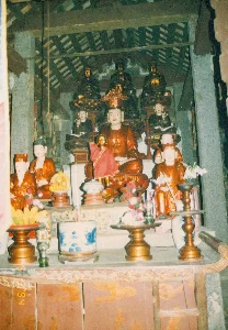 Đây là khung cảnh bên trong một ngôi chùa hoặc đền thờ ở khu vực Ninh Bình, Tam Cốc. Bức ảnh chụp một bàn thờ Phật được bài trí công phu và trang nghiêm.。Bàn thờ được chia thành nhiều tầng. Tầng trên cùng có các pho tượng Phật bằng đồng hoặc sơn mài sẫm màu, ngồi trên đài sen. Tầng giữa, ở vị trí trung tâm, là một pho tượng lớn, có thể là tượng Phật hoặc Bồ tát, mặc áo cà sa màu cam và đội mũ miện. Phía trước tượng chính có một pho tượng nhỏ hơn, mạ vàng, mặc áo đỏ. Xung quanh là nhiều pho tượng nhỏ hơn với các tư thế và trang phục khác nhau, được đặt trên các bệ thờ.。Trên bàn thờ phía trước, có nhiều đồ lễ cúng được bày biện. Nổi bật là một lư hương lớn bằng sứ men lam trắng, bên trong có cắm hương (nhang). Ngoài ra còn có các mâm bồng bằng gỗ đựng hoa quả (chuối), các bình hoa cắm hoa tươi như hoa loa kèn trắng và hoa hồng, cùng với các chân đèn dầu.。Kiến trúc của không gian thờ cúng có các cột trụ bằng đá và kết cấu mái gỗ với ngói lợp lộ ra bên trong. Bàn thờ được làm bằng gỗ, với các tấm ván được trang trí hoa văn. Ở góc dưới bên trái của bàn thờ có thể thấy con số "'94", cho biết thời điểm chụp ảnh là năm 1994. Khung cảnh cho thấy một không gian tín ngưỡng Phật giáo truyền thống của Việt Nam.。（日本語訳）。これはニンビン、タムコック地域にある寺院または祠堂の内部の光景です。写真は、精巧かつ荘厳に飾られた仏壇を写しています。仏壇は多層に分かれています。最上段には、蓮台に座った、銅製または暗色の漆塗りの仏像があります。中段の中央には、オレンジ色の袈裟をまとい、宝冠をかぶった、おそらく仏陀または菩薩の大きな像があります。主像の前には、金メッキが施され、赤い衣を着た、より小さな像があります。その周りには、様々な姿勢や装束の、より小さな像が多数、祭壇の上に置かれています。手前の祭壇の上には、多くのお供え物が並べられています。目立つのは、中に線香が立てられた、青と白の磁器でできた大きな香炉です。その他にも、果物（バナナ）を盛った木製の高坏（たかつき）、白いユリやバラなどの生花が生けられた花瓶、そして油灯の燭台があります。仏壇は木製で、板には模様が装飾されています。仏壇の左下隅には「'94」という数字が見え、撮影時期が1994年であることを示しています。この光景は、ベトナムの伝統的な仏教信仰の空間を示しています。