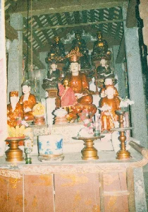 Bức ảnh chụp một bàn thờ trong một ngôi chùa hoặc đền thờ, có thể là ở khu vực Ninh Bình, Tam Cốc. Đây là một không gian thờ cúng tôn giáo trang nghiêm. Bàn thờ được sắp xếp theo nhiều tầng bậc. Ở vị trí trung tâm là một pho tượng lớn, có thể là Phật hoặc Bồ tát, mặc áo choàng màu cam và đội mũ miện. Xung quanh và ở các tầng cao hơn là nhiều pho tượng khác nhỏ hơn, một số được sơn son thếp vàng, ngồi trong tư thế thiền định trên đài sen. Phía trước tượng chính có một bức tượng nhỏ hơn đang đứng trong trang phục màu đỏ. Trên bàn thờ phía trước bày các vật phẩm cúng dường. Có thể thấy rõ một bát hương lớn bằng sứ hoa lam, các đĩa gỗ (mâm bồng) đựng hoa quả như chuối, cùng với các bình hoa tươi cắm hoa loa kèn trắng và hoa hồng. Ngoài ra còn có các chân nến và đèn dầu. Kiến trúc xung quanh cho thấy đây là một công trình cổ, với các cột trụ bằng đá và mái nhà có kết cấu dầm gỗ lộ ra. Ở phía bên trái có treo một dải giấy màu đỏ có chữ viết nhưng không thể đọc được. Các tấm gỗ ở chân bàn thờ cũng có các hình vẽ trang trí nhưng đã bị mờ theo thời gian.。（日本語訳）。この写真は、おそらくニンビンやタムコック地域にある寺院または祠堂の祭壇を撮影したものです。これは荘厳な宗教的礼拝空間です。祭壇は多層に配置されています。中央には、オレンジ色の袈裟をまとい、宝冠をかぶった、おそらく仏陀または菩薩の大きな像があります。その周りや上段には、より小さな他の多くの像があり、そのいくつかは金箔で朱塗りされ、蓮華座の上で瞑想の姿勢で座っています。主像の前には、赤い衣装を着た、より小さな立像があります。手前の祭壇には供物が置かれています。青と白の磁器でできた大きな香炉、バナナなどの果物を盛った木製の皿（高坏）、白いユリやバラを生けた花瓶がはっきりと見えます。その他に、燭台や油灯もあります。周りの建築は、石の柱と木製の梁が露出した屋根構造を持つ、古い建物であることを示しています。左側には文字が書かれた赤い紙が吊るされていますが、読むことはできません。祭壇の足元にある木の板にも装飾的な絵がありますが、時間とともに色褪せています。