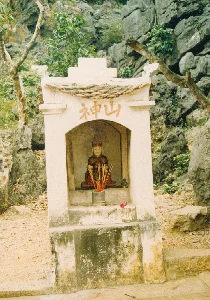 Bức ảnh chụp một am thờ nhỏ được dựng ngoài trời, dưới chân một vách núi đá. Phía trên khám thờ có ghi hai chữ Hán màu đỏ là '神山' (Thần Sơn), có nghĩa là 'Núi Thiêng'. Bên trong am thờ có đặt một bức tượng của một vị thần, mặc trang phục và đội mũ kiểu cổ. Phía trước tượng là một bát hương bằng kim loại, bên trong cắm rất nhiều chân hương đã cháy, cho thấy hoạt động thờ cúng đã diễn ra. Ngoài ra còn có một chiếc đèn dầu nhỏ và một bông hoa được đặt trên bệ thờ. Toàn bộ am thờ có vẻ cũ, phần chân đế bị rêu phong, bám bẩn. Khung cảnh xung quanh là núi đá và cây cối, phù hợp với địa hình của khu vực Tam Cốc, Ninh Bình.。（日本語訳）。写真には、岩壁の麓、屋外に建てられた小さな祠が写っている。祠の上部には赤い漢字で「神山」（Thần Sơn）と書かれており、「聖なる山」を意味する。像の前には金属製の香炉があり、中には燃え尽きた線香の芯がたくさん刺さっており、礼拝が行われたことを示している。祠全体は古びて見え、土台部分は苔むし、汚れている。周囲の風景は岩山と木々であり、ニンビン省タムコック地域の地形と一致している。