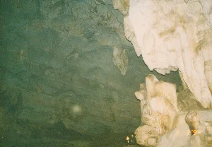 Bên trong một hang động, có thể là một trong các hang động ở Tam Cốc, Ninh Bình. Trần và vách hang có màu xanh xám với các lớp đá ngang. Phía bên phải là một khối thạch nhũ lớn màu trắng ngà, trong khi các khối măng đá nhỏ hơn nằm ở phía dưới. Có nhiều thạch nhũ với các hình dạng khác nhau treo từ trần hang. Ở phía dưới, hai chiếc đèn dầu nhỏ được đặt trên một mỏm đá, đang cháy và tỏa ra ánh sáng vàng để chiếu sáng không gian. Có thể nhìn thấy một vài chữ viết hoặc hình vẽ mờ trên khối đá màu trắng nhưng không thể đọc được nội dung. Khung cảnh cho thấy đây là một điểm tham quan tự nhiên được khai thác cho du lịch vào thời điểm đó.。（日本語訳）。洞窟の中、おそらくニンビン省タムコックにある洞窟の一つだろう。洞窟の天井と壁は、水平な岩の層を持つ青灰色をしている。右側には象牙色の大きな鍾乳石の塊があり、その下にはより小さな石筍の塊がある。洞窟の天井からは、さまざまな形の鍾乳石が多数垂れ下がっている。下の方では、岩棚の上に2つの小さなオイルランプが置かれ、燃えて黄色い光を放ち、空間を照らしている。この光景は、ここが当時、観光用に開発された自然の観光地であったことを示している。