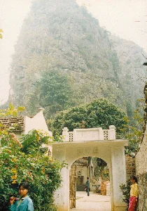 Bối cảnh là một khung cảnh làng quê ven sông ở vùng Ninh Bình, Tam Cốc vào năm 1994. Dưới lòng sông, có nhiều hoạt động vận chuyển bằng thuyền. Nổi bật là một chiếc thuyền tam bản nhỏ có gắn buồm, trên đó một người phụ nữ đang dùng sào đẩy thuyền đi trong khi một đứa trẻ ngồi phía trước. Một chiếc thuyền khác lớn hơn, có mái che, đang chở đầy đá, cho thấy hoạt động vận chuyển vật liệu xây dựng. Một chiếc thuyền thứ ba neo đậu gần bờ, bên cạnh là các đống gạch và vật liệu xây dựng khác. Trên bờ là những ngôi nhà cấp bốn lợp ngói, một số nhà đang trong quá trình xây dựng với tường gạch xi măng chưa trát. Có một vài người dân trên bờ và cây cối xanh tươi. Phía xa, những dãy núi đá vôi đặc trưng của khu vực ẩn hiện trong sương mù. Khung cảnh phản ánh cuộc sống lao động và sinh hoạt bình dị của người dân vùng sông nước Bắc Bộ.。（日本語訳）。舞台は1994年、ニンビン省タムコック地方の川沿いの村の風景です。川面では、船による輸送活動が盛んに行われています。目立っているのは、帆を付けた小さな三板船（サンパン）で、その上では一人の女性が竿を使って船を押し進めており、前には一人の子供が座っています。もう一艘のより大きな屋根付きの船は、石を満載しており、建設資材の輸送活動を示しています。三艘目の船は岸の近くに停泊しており、その傍らにはレンガやその他の建設資材の山があります。岸には瓦葺きの平屋があり、いくつかの家はまだ漆喰が塗られていないセメントレンガの壁で、建設中です。岸には数人の住民と青々とした木々が見られます。遠くには、この地域特有の石灰岩の山々が霧の中に姿を現したり隠したりしています。この風景は、北部水郷地帯の人々の素朴な労働と日常生活を反映しています。