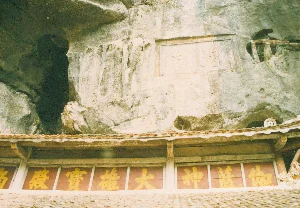 Bức ảnh chụp một ngôi chùa được xây dựng dựa vào vách núi đá vôi, có khả năng là ở khu vực Tam Cốc, Ninh Bình. Cảnh quan cho thấy một hang động tối ở phía bên trái và các chữ Hán được khắc trực tiếp lên vách đá. Một tấm bia đá hình chữ nhật khắc hai chữ có thể là "仙洞" (Tiên Động). Bên dưới vách đá là mái hiên lợp ngói của ngôi chùa. Ngay dưới mái hiên là một bức hoành phi dài màu nâu đỏ với các chữ Hán lớn màu vàng. Các chữ này đọc từ phải sang trái bao gồm "大雄寶殿" (Đại Hùng Bảo Điện), là tên gọi của chính điện trong chùa Phật giáo, và "伽藍神" (Già Lam Thần), chỉ các vị thần hộ pháp. Toàn bộ khung cảnh thể hiện sự kết hợp giữa kiến trúc tôn giáo cổ và cảnh quan thiên nhiên đặc trưng của khu vực.。（日本語訳）。写真は、石灰岩の崖に寄り添うように建てられた寺院を写しており、おそらくニンビン省タムコック地域にあるものと思われます。風景には左側に暗い洞窟が見え、崖には漢字が直接刻まれています。長方形の石碑には、「仙洞」（ティエン・ドン）である可能性のある二文字が刻まれています。崖の下には、寺院の瓦葺きの軒があります。軒のすぐ下には、大きな黄色い漢字が書かれた赤褐色の長い扁額があります。これらの文字は右から左に読み、仏教寺院の本堂の名称である「大雄寶殿」（ダイフンバオディエン）と、護法神を指す「伽藍神」（ザーラムタン）が含まれています。全体の光景は、古代の宗教建築とこの地域特有の自然景観との融合を表しています。