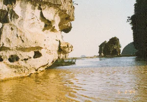 Bức ảnh chụp phong cảnh tại khu vực Tam Cốc, Ninh Bình. Khung cảnh chủ đạo là một dòng sông có màu nước vàng đục, chảy giữa những dãy núi đá vôi hùng vĩ. Phía bên trái là một vách núi đá lớn với bề mặt bị bào mòn, tạo thành nhiều hốc và hình thù độc đáo. Cây cối mọc rải rác trên các vách núi. Ở phía xa, có thể nhìn thấy một cây cầu thấp hoặc một con đường nhỏ bắc ngang qua sông. Toàn cảnh mang vẻ đẹp hoang sơ, yên bình. Góc chụp cho thấy khả năng cao bức ảnh được chụp từ một chiếc thuyền đang du ngoạn trên sông. Trên mặt nước ở góc dưới bên phải có dấu ngày tháng kỹ thuật số màu cam '5 2 '94', tương ứng với ngày 5 tháng 2 năm 1994.。（日本語訳）。ニンビン省、タムコック地域の風景を撮影した写真。主な風景は、雄大な石灰岩の山々の間を流れる、黄色く濁った水色の川です。左側には大きな岩壁があり、その表面は侵食され、多くのくぼみや独特の形状を作り出しています。崖の上には木々がまばらに生えています。遠くには、川を横切る低い橋か小さな道が見えます。全景は、自然のままの穏やかな美しさを醸し出しています。撮影角度から、この写真は川を遊覧中のボートから撮影された可能性が高いことがわかります。右下の水面には、1994年2月5日に相当するオレンジ色のデジタル日付スタンプ「'5 2 '94」があります。