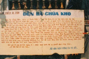 Bối cảnh là một lễ hội rất đông người đang diễn ra tại một khu vực đền chùa. Một đám đông lớn gồm đàn ông, phụ nữ và trẻ em đang tụ tập trên các bậc thang bằng đá và gạch. Phía sau họ là một cổng chùa được sơn màu vàng, trên đó có dòng chữ viết tay màu đỏ: "Kính chào qúi khách VỀ TƯỞNG NIỆM BÀ CHÚA KHO". Trên các cột của cổng có các dòng chữ Hán nhưng không thể đọc rõ. Phía trên cổng có cắm một lá cờ đỏ sao vàng và một chiếc loa phóng thanh. Nhiều lá cờ lễ hội khác cũng được treo xung quanh. Mọi người đang tham dự lễ hội, một số ngồi trên các bậc thang, một số đứng xem. Có người che ô màu hồng và ô nhiều màu để tránh nắng. Một người phụ nữ ở phía trước đang bưng một mâm lễ vật. Trang phục của mọi người phản ánh phong cách thời trang năm 1994. Khung cảnh chung cho thấy một không khí lễ hội náo nhiệt tại một di tích lịch sử.。（日本語訳）。背景は、寺社仏閣のある地域で開かれている、非常に多くの人で賑わう祭りです。男性、女性、子供を含む大群衆が、石とレンガでできた階段の上に集まっています。彼らの後ろには黄色く塗られた寺の門があり、その上には「バー・チュア・コー夫人追悼にお越しの皆様を歓迎いたします」という赤い手書きの文字があります。門の柱には漢字の文字列がありますが、はっきりと読むことはできません。門の上には、金星紅旗が一本と拡声器が一つ設置されています。他の多くの祭りの旗も周りに掲げられています。人々は祭りに参加しており、階段に座っている人もいれば、立って見物している人もいます。日差しを避けるために、ピンク色の傘や多色の傘をさしている人がいます。前方の一人の女性が、お供え物を載せたお盆を運んでいます。人々の服装は1994年のファッションスタイルを反映しています。全体的な光景は、歴史的遺跡での賑やかな祭りの雰囲気を示しています。