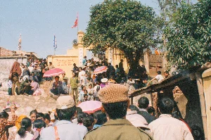 Bức ảnh ghi lại cảnh một đám đông người đang tham gia một nghi lễ tôn giáo tại một nơi có vẻ là chùa hoặc đền vào ngày 19 tháng 2 năm 1994. Không khí nghi ngút khói hương, cho thấy hoạt động thắp hương đang diễn ra. Ở trung tâm, một người phụ nữ đang chắp tay thành kính, có lẽ đang cầu nguyện trước một ban thờ với hoa và các lễ vật khác. Ở phía trước bên trái, một người đang bưng một mâm quả cúng gồm những quả cam. Xung quanh có nhiều người khác, bao gồm một người đàn ông mặc vest và một người đàn ông đội mũ lưỡi trai ở tiền cảnh. Phía sau có thể thấy một bức tường gạch và một tấm bạt trắng được căng lên. Trên ảnh có dấu ngày tháng kỹ thuật số màu cam ghi '19 2 '94'.。（日本語訳）。1994年2月19日、寺か廟と思われる場所で宗教儀式に参加している人々の群衆の光景を記録した写真。線香の煙が立ち込め、線香を焚く活動が行われていることを示している。中央では、一人の女性が敬虔に手を合わせ、おそらく花やその他のお供え物がある祭壇の前で祈っている。左手前では、一人の人がオレンジを含む果物のお供え物の盆を運んでいる。周りには他の多くの人々がおり、前景にはベストを着た男性と野球帽をかぶった男性が含まれている。写真には「19 2 '94」と記されたオレンジ色のデジタル日付スタンプがある。
