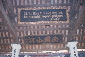 Bối cảnh là bên trong một công trình kiến trúc cổ, có thể là một ngôi đình làng hoặc đền thờ ở khu vực Phú Thọ. Mái nhà lợp ngói đất nung với kết cấu vì kèo và xà nhà bằng gỗ.。Tâm điểm của bức ảnh là một chiếc trống lớn truyền thống, được treo trên xà nhà. Mặt trống màu trắng, thân trống màu đỏ. Trên mặt trống có ghi dòng chữ: "TU SỬA TẠI CỬA HÀNG PHÚ LỘC HUYỆN PHONG CHÂU .1993.". Dòng chữ này cho biết chiếc trống được tu sửa vào năm 1993 tại một cửa hàng ở Phú Lộc, huyện Phong Châu (một huyện cũ nay thuộc tỉnh Phú Thọ).。Phía sau chiếc trống, có một tấm bảng lớn màu đỏ khắc rất nhiều chữ, nhưng không thể đọc rõ nội dung, có thể là bảng công đức hoặc văn bia ghi lại lịch sử. Có nhiều người đang ở trong không gian này, dường như là khách tham quan. Một người đàn ông mặc áo khoác xanh, quần jean, đeo kính và tay cầm máy ảnh. Xung quanh còn có những người khác, bao gồm một cô gái tết tóc đang đi ngang qua. Một chiếc bàn gỗ đơn sơ được kê gần đó, trên bàn có vài hộp đồ. Khung cảnh cho thấy một chuyến tham quan di tích văn hóa, lịch sử vào thời điểm năm 1994.。（日本語訳）。背景は、フートー省地域にある古い建築物、おそらく村の亭（ディン）か祠の内部です。屋根は素焼きの瓦で葺かれ、木製の垂木と梁の構造になっています。写真の中心は、梁に吊るされた大きな伝統的な太鼓です。太鼓の面は白く、胴体は赤いです。太鼓の面には「TU SỬA TẠI CỬA HÀNG PHÚ LỘC HUYỆN PHONG CHÂU .1993.」という文字が書かれています。この文字は、この太鼓が1993年にフォンチャウ県（現在はフートー省に属する旧県）のフーロックにある店で修理されたことを示しています。太鼓の後ろには、多くの文字が刻まれた大きな赤い板がありますが、内容ははっきりと読み取れず、功徳碑か歴史を記録した石碑かもしれません。この空間には多くの人がおり、観光客のようです。一人の男性は青いジャケットを着てジーンズを履き、眼鏡をかけ、手にカメラを持っています。周りには、通り過ぎていく三つ編みの女の子を含め、他の人々もいます。近くには簡素な木製の机が置かれており、その上にはいくつかの箱があります。この光景は、1994年当時の文化歴史遺跡の見学の様子を示しています。