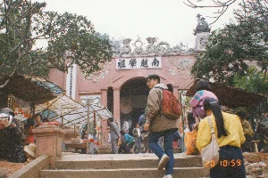 Trong ảnh là một bé gái đang ngồi ở ngưỡng cửa của một tòa nhà, có thể là một ngôi chùa hoặc một ngôi nhà. Cô bé mặc một chiếc áo dài tay màu hồng có hoa văn, quần tối màu và quàng một chiếc khăn len màu vàng quanh cổ. Trên tay cô bé đang ôm một bó hương (nhang) lớn màu hồng, loại thường dùng để đi lễ. Các que hương có dán nhãn giấy nhiều màu sắc nhưng không thể đọc được chữ. Khung cửa bằng gỗ sơn màu đỏ đã cũ và bong tróc, bức tường bên cạnh màu trắng cũng có dấu hiệu của thời gian với lớp vữa bị tróc. Không gian bên trong tối, không thể nhìn rõ chi tiết. Cảnh tượng này mô tả một khoảnh khắc đời thường, có thể liên quan đến hoạt động tín ngưỡng tại một địa điểm ở miền Bắc Việt Nam vào khoảng năm 1994.。（日本語訳）。写真には、ある建物（寺院か家かもしれない）の戸口に座っている一人の女の子が写っている。女の子は模様のあるピンク色の長袖の服、暗い色のズボンを履き、首には黄色い毛糸のスカーフを巻いている。手には、お参りによく使われる種類の、大きなピンク色の線香の束を抱えている。線香には色とりどりの紙ラベルが貼られているが、文字は読めない。赤く塗られた木製の戸枠は古びて剥がれており、隣の白い壁も漆喰が剥がれ、時の経過の痕跡が見られる。中の空間は暗く、詳細をはっきりと見ることはできない。この光景は、1994年頃の北ベトナムのある場所での信仰活動に関連する可能性のある、日常の一瞬を描写している。