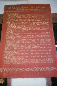 Bức ảnh chụp cận cảnh một tấm bảng lớn màu đỏ với chữ khắc màu vàng, được gọi là "Bảng vàng ghi công", đặt tại khu di tích lịch sử Đền Hùng, tỉnh Phú Thọ. Tấm bảng này ghi lại danh sách các cá nhân, tổ chức và địa phương đã đóng góp công đức để tu bổ và xây dựng Đền Hùng qua nhiều thời kỳ, từ năm 1912 đến năm 1990. Đây là một hiện vật lịch sử, thể hiện sự đóng góp của người dân cả nước và kiều bào ở nước ngoài đối với việc bảo tồn di tích.。Nội dung chi tiết trên bảng được ghi lại như sau:。BẢNG VÀNG GHI CÔNG。TU BỔ ĐỀN HÙNG。NĂM 1912 - 1922:。- 18 TỈNH MIỀN BẮC ỦNG HỘ 6.000,00 ĐỒNG.。- BÀ NGUYỄN THỊ GAI (ĐÔNG XUÂN - HÀ NỘI) ỦNG HỘ 1.000,00 ĐỒNG, LÀM ĐƯỜNG TỪ CỔNG ĐỀN LÊN ĐỀN THƯỢNG.。- HIỆU ĐÔNG THỊNH (HÀ NỘI) ỦNG HỘ XÂY DỰNG CỔNG ĐỀN.。NĂM 1981:。- BÀ NGUYỄN THỊ MẠCH (TÂN HÙNG, BA VÌ, HÀ SƠN BÌNH) CÙNG MỘT SỐ ĐỒNG BÀO CUNG TIẾN 20.000,00 ĐỒNG.。NĂM 1983:。- TỈNH TIỀN GIANG CUNG TIẾN TỦ THỜ BẰNG GỖ CẨM LAI.。- TỈNH ĐỒNG NAI CUNG TIẾN HAI LỌ LỤC BÌNH GỐM QUÝ.。- CÁC CỤ PHỤ LÃO THÀNH PHỐ HỒ CHÍ MINH CUNG TIẾN HAI CÂY SÁP RỒNG, 19 CÂY HƯƠNG LỚN.。- SỞ VĂN HÓA THÔNG TIN QUẢNG NAM - ĐÀ NẴNG CUNG TIẾN HAI NỒI NHANG BẰNG ĐÁ MÀU NGŨ HÀNH SƠN.。- BÀ PHÙNG THỊ QUÝ (ĐƯỜNG ĐÀ NẴNG HẢI PHÒNG) ỦNG HỘ 2.000,00 ĐỒNG.。NĂM 1984:。- UBND TỈNH BẾN TRE CUNG TIẾN BỘ ĐỈNH BẰNG ĐỒNG.。NĂM 1986 - 1989:。- THÀNH ỦY - UBND - MẶT TRẬN TỔ QUỐC THÀNH PHỐ HÀ NỘI CUNG TIẾN 10 TRIỆU ĐỒNG VÀ TƯỢNG BÁC HỒ BẰNG ĐỒNG.。- UBND TỈNH AN GIANG CUNG TIẾN 10 TRIỆU ĐỒNG.。- NHÂN DÂN NGHỆ TĨNH CUNG TIẾN 5 TRIỆU ĐỒNG.。- HỘI ĐỒNG HƯƠNG VĨNH PHÚ TẠI HÀ NỘI CUNG TIẾN 650.000 ĐỒNG.。- BÀ NGUYỄN THỊ TRUYỀN VIỆT KIỀU TẠI MỸ CUNG TIẾN MỘT BỘ TRỐNG LỚN TẠI ĐỀN THƯỢNG.。- ÔNG VŨ TIẾN THANH VIỆT KIỀU Ở NHẬT CUNG TIẾN 300 ĐÔ LA.。- ÔNG NGUYỄN XUÂN HỒI VIỆT KIỀU Ở PHÁP CUNG TIẾN 100.000 ĐỒNG.。- NHÂN DÂN THẬP PHƯƠNG ỦNG HỘ 6 TRIỆU ĐỒNG.。NĂM 1990:。- BỘ VĂN HÓA CẤP 20 TRIỆU ĐỒNG MỞ RỘNG SÂN LĂNG HÙNG VƯƠNG.。- UBND TỈNH VĨNH PHÚ CẤP TRÊN 500 TRIỆU ĐỒNG ĐỂ MỞ ĐƯỜNG TỪ LĂNG XUỐNG ĐỀN GIẾNG, HOÀN THIỆN PHẦN XÂY LẮP NHÀ BẢO TÀNG HÙNG VƯƠNG.。（日本語訳）。この銘板には、1912年から1990年までの様々な時代にわたり、フン王の祠の修復と建設のために功徳を寄進した個人、組織、地方のリストが記録されている。これは、遺跡の保存に対する全国民および海外在住ベトナム人同胞の貢献を示す歴史的な遺物である。銘板の詳細な内容は以下の通り記録されている。功績を記録する黄金の銘板。フン王の祠の修復。1912年 - 1922年：。- 北部18省が6,000.00ドンを寄付。- グエン・ティ・ガイ女史（ハノイ市ドン・スアン）が1,000.00ドンを寄付し、祠の門から上祠までの道を整備。- ドン・ティン商号（ハノイ市）が祠の門の建設を支援。1981年：。- グエン・ティ・マック女史（ハソンビン省バビ県タンフン）が一部の同胞と共に20,000.00ドンを奉納。1983年：。- ティエンザン省が紫檀製の祭壇を奉納。- ドンナイ省が貴重な陶器製の六角瓶二つを奉納。- ホーチミン市の長老たちが龍の蝋燭二本、大きな線香19本を奉納。- クアンナム・ダナン文化情報局が五行山の色の石で作られた香炉二つを奉納。- フン・ティ・クイ女史（ハイフォン市ダナン通り）が2,000.00ドンを寄付。1984年：。- ベンチェ省人民委員会が銅製の香炉一式を奉納。1986年 - 1989年：。- ハノイ市党委員会 - 人民委員会 - 祖国戦線が1,000万ドンと銅製のホーおじさんの像を奉納。- アンザン省人民委員会が1,000万ドンを奉納。- ゲティン省の人民が500万ドンを奉納。- ハノイ在住ヴィンフー省出身者会が65万ドンを奉納。- 在米ベトナム人同胞のグエン・ティ・チュエン女史が上祠に大きな太鼓一式を奉納。- 在日ベトナム人同胞のヴー・ティエン・タイン氏が300ドルを奉納。- 在仏ベトナム人同胞のグエン・スアン・ホイ氏が10万ドンを奉納。- 十方の人民が600万ドンを寄付。1990年：。- 文化省がフン王陵の庭の拡張のために2,000万ドンを支給。- ヴィンフー省人民委員会が陵から井戸の祠までの道を開通し、フン王博物館の建設・設置部分を完成させるために5億ドン以上を支給。