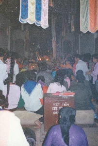 Trên một hòm công đức bằng gỗ đặt ở phía trước có ghi dòng chữ: 'KÉT TIỀN CUNG TIẾN ĐỀN'. Bối cảnh là không gian bên trong một ngôi đền vào buổi tối, rất đông người dân đang tập trung tham dự một nghi lễ hoặc lễ hội. Mọi người ngồi trên sàn và các bệ cao, hướng mặt về phía ban thờ chính ở sâu bên trong, nơi có ánh sáng của nến và đèn hương. Trên trần nhà được trang trí bằng nhiều dải cờ phướn lễ hội với các màu sắc sặc sỡ như xanh, trắng, hồng, cam. Nhiều phụ nữ trong ảnh để tóc dài, một số tết tóc hoặc mặc trang phục truyền thống. Khung cảnh phản ánh một sinh hoạt tín ngưỡng cộng đồng trang nghiêm tại một ngôi đền ở Việt Nam vào năm 1994.。（日本語訳）。前方に置かれた木製の賽銭箱には、「KÉT TIỀN CUNG TIẾN ĐỀN」（寺院への奉納金庫）という文字が書かれている。場面は夜のお寺の内部で、非常に多くの人々が儀式か祭りに参加するために集まっている。人々は床や高い台座の上に座り、奥深くにある、ろうそくや線香の灯りがある本堂の祭壇の方を向いている。天井は、青、白、ピンク、オレンジといった色とりどりの祭りの幟旗で飾られている。写真に写っている多くの女性は髪が長く、一部は髪を結っていたり、伝統的な衣装を着ていたりする。この光景は、1994年のベトナムのある寺院における、厳かな共同体の信仰活動を反映している。