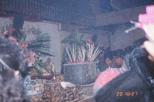 Bối cảnh là bên trong một ngôi đình, chùa hoặc một nơi thờ cúng tâm linh, có kiến trúc dầm gỗ và trần nhà được trang trí. Một đám đông đang tụ tập trang nghiêm xung quanh một bàn thờ lớn để thực hiện nghi lễ cúng bái. Trên bàn thờ có một lư hương lớn bằng kim loại hoặc đá, được chạm khắc hoa văn tinh xảo, bên trong cắm đầy nhang đang cháy và tỏa khói nghi ngút. Xung quanh lư hương là các lễ vật như hoa tươi (hoa cúc, hoa đồng tiền) cắm trong lọ sứ men lam trắng, trái cây (cam, quýt), và một ngọn nến đang cháy. Phía sau bàn thờ, trên tường có các bức vẽ, trong đó có thể nhận ra hình đầu rồng. Mọi người đều hướng về phía bàn thờ với vẻ thành kính. Đây có thể là một hoạt động trong một lễ hội đầu năm, vì thời điểm chụp là ngày 20 tháng 2 năm 1994, ngay sau Tết Nguyên Đán. Trên hình ảnh có dấu ngày tháng của máy ảnh ghi '20 10:27'.。（日本語訳）。背景は、お堂、寺院、または何らかの精神的な礼拝所の内部で、木製の梁と装飾された天井を持つ建築様式です。大勢の人々が、礼拝の儀式を行うために大きな祭壇の周りに厳かに集まっています。祭壇の上には、金属または石でできた大きな香炉があり、精巧な模様が彫刻され、中には燃えている線香がたくさん立てられ、煙がもうもうと立ち上っています。香炉の周りには、青と白の磁器の花瓶に生けられた生花（菊、ガーベラ）、果物（オレンジ、みかん）、そして燃えているろうそく一本といったお供え物があります。祭壇の後ろの壁には絵があり、その中には龍の頭の形が見て取れます。皆、敬虔な面持ちで祭壇の方を向いています。これは年始の祭りの一環の活動である可能性があります。なぜなら、撮影時期は1994年2月20日で、旧正月（テト）の直後だからです。画像には、カメラの日付スタンプで「20 10:27」と記録されています。