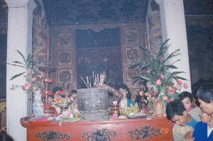 Bức ảnh ghi lại khung cảnh bên trong một ngôi đền hoặc đình cổ ở Việt Nam, có thể là trong một dịp lễ hội đầu xuân vào khoảng năm 1994. Không gian rất đông đúc, trang nghiêm với nhiều người dân đang thành kính cúng bái.。Ở vị trí trung tâm là một bàn thờ lớn bằng gỗ sơn màu đỏ, được chạm khắc các họa tiết màu đen tinh xảo. Trên bàn thờ, một lư hương lớn bằng đá hoặc kim loại đang cắm đầy nhang (hương) khói nghi ngút. Xung quanh lư hương là các lễ vật được người dân dâng cúng, bao gồm nhiều loại hoa quả như chuối, cam, quýt; hai bình hoa lớn cắm hoa đồng tiền và các loại hoa khác; cùng với vàng mã và các đồ lễ khác.。Phía sau bàn thờ là một gian thờ với các cánh cửa gỗ được chạm trổ phức tạp. Phía trên gian thờ có một bức hoành phi lớn, trên đó có khắc ba chữ Hán/Nôm lớn nhưng khó đọc rõ toàn bộ nội dung do chất lượng ảnh và góc chụp. Có thể nhận ra một vài nét của các chữ như '王' (Vương) và '基' (Cơ). Hai bên là các cột trụ lớn màu trắng, và trên vách tường bên cạnh cột có thể thấy các hình vẽ trang trí nhiều màu sắc, dường như là hình rồng.。Mọi người, từ người già đến thanh niên, đang tụ tập xung quanh bàn thờ để dâng hương và cầu nguyện, tạo nên một không khí tâm linh đặc trưng của các lễ hội truyền thống Việt Nam.。（日本語訳）。この写真は、1994年頃の初春の祭りの際に撮影されたと思われる、ベトナムの古い寺院または亭（ディン）の内部の光景を記録したものです。空間は非常に混雑しており、多くの人々が敬虔にお参りをしていて、厳かな雰囲気です。中央には、赤く塗られた木製の大きな祭壇があり、黒い精巧な模様が彫刻されています。祭壇の上には、石または金属製の大きな香炉があり、線香がびっしりと立てられ、煙がもうもうと立ち上っています。香炉の周りには、人々がお供えした供物があり、バナナ、オレンジ、ミカンなどの多くの種類の果物、ガーベラやその他の花が生けられた二つの大きな花瓶、そして冥銭やその他の儀式用品が含まれています。祭壇の後ろには、複雑な彫刻が施された木製の扉を持つ祠があります。祠の上には大きな扁額があり、そこには3つの大きな漢字/チュノムが刻まれていますが、写真の画質と撮影角度のため、内容全体をはっきりと読み取ることは困難です。「王」（Vương）や「基」（Cơ）といった文字のいくつかの筆画を認識することができます。両側には大きな白い柱があり、柱の隣の壁には、龍の絵と思われる色鮮やかな装飾画が見られます。老人から若者まで、あらゆる人々が祭壇の周りに集まり、線香をあげて祈りを捧げており、ベトナムの伝統的な祭りに特有の精神的な雰囲気を醸し出しています。
