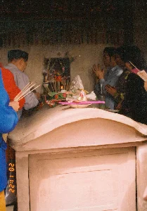 Trong ảnh có dấu thời gian của máy ảnh ghi '22:11 02'. Ở phía sau, trên tường, có các họa tiết trang trí và có thể có chữ viết nhưng không thể đọc rõ.。Bối cảnh là một không gian thờ cúng trong nhà, có thể là một lăng mộ hoặc nhà thờ họ. Ở trung tâm là một ngôi mộ lớn bằng xi măng với nắp hình vòm. Một nhóm người đang thực hiện nghi lễ cúng bái. Một người đàn ông lớn tuổi đội mũ đang đứng trước ngôi mộ, thực hiện các nghi thức. Một người khác đang cầm một bó hương lớn chuẩn bị thắp. Ở phía bên phải, những người khác đang chắp tay thành kính khấn vái.。Trên nắp ngôi mộ được dùng làm bàn thờ, có bày các lễ vật cúng bái bao gồm hoa, một cây nến đang cháy, hương đang cháy trong lư, và các đồ cúng khác. Đây là một nghi lễ thờ cúng tổ tiên hoặc một vị thần, một hoạt động tín ngưỡng truyền thống ở Việt Nam.。（日本語訳）。写真には「22:11 02」と記録されたカメラのタイムスタンプがあります。背景は屋内の祭祀空間で、霊廟または一族の祠堂である可能性があります。中央には、アーチ型の蓋が付いたセメント製の大きな墓があります。一団の人々が礼拝の儀式を行っています。帽子をかぶった年配の男性が墓の前に立ち、儀式を執り行っています。別の人は、火をつける準備をしながら大きな線香の束を持っています。右側では、他の人々が敬虔に手を合わせて祈っています。祭壇として使われている墓の蓋の上には、花、燃えているろうそく、香炉で燃えている線香、その他の供物を含む礼拝用のお供え物が並べられています。これは祖先または神を祀る儀式であり、ベトナムの伝統的な信仰活動です。