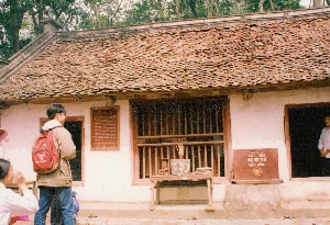 Bản ghi chữ viết: Trên tường có treo một tấm biển gỗ, dòng trên cùng ghi 'ĐỀN TRUNG', phần văn bản bên dưới không thể đọc rõ nhưng có khả năng là thông tin về lịch sử ngôi đền. Bên cạnh đó là một hòm công đức bằng gỗ màu nâu đỏ với dòng chữ trắng 'KÉT TIỀN CUNG TIẾN TU BỔ ĐỀN HÙNG'.。Mô tả cảnh quan và hoạt động: Bối cảnh là phía trước một ngôi đền cổ, có khả năng là Đền Trung trong Khu di tích lịch sử Đền Hùng, tỉnh Phú Thọ. Ngôi đền có kiến trúc truyền thống Việt Nam với mái lợp ngói âm dương đã cũ, tường quét vôi trắng và cửa gỗ. Phía sau đền là những cây cổ thụ. Một nam thanh niên mặc áo khoác, đeo ba lô màu đỏ đang đứng xem tấm biển thông tin treo trên tường. Phía trước cửa sổ có đặt một chiếc bàn gỗ nhỏ làm bàn thờ tạm, trên bàn có một bát hương bằng sứ cắm đầy nhang (hương) và một số vật phẩm thờ cúng khác. Hoạt động chính là mọi người đến thăm viếng và dâng hương tại di tích lịch sử.。（日本語訳）。書かれた文字の記録：壁には木製の看板が掛けられており、一番上の行には「ĐỀN TRUNG」（チュン祠）と書かれている。その下のテキスト部分ははっきりと読むことはできないが、おそらく祠の歴史に関する情報であろう。その隣には赤褐色の木製のお賽銭箱があり、白い文字で「KÉT TIỀN CUNG TIẾN TU BỔ ĐỀN HÙNG」（フン祠修復のための奉納金庫）と書かれている。景観と活動の説明：背景は古い祠の前で、おそらくフート省のフン祠歴史遺跡地区内にあるチュン祠であろう。この祠はベトナムの伝統的な建築様式で、古くなった陰陽瓦で葺かれた屋根、白い漆喰で塗られた壁、そして木製の扉がある。祠の後ろには古い木々がある。ジャケットを着て、赤いバックパックを背負った一人の若い男性が、壁に掛けられた案内板を見ている。窓の前には仮の祭壇として小さな木製のテーブルが置かれており、テーブルの上には線香がいっぱいに立てられた陶器の香炉と、その他のいくつかのお供え物がある。主な活動は、人々がこの歴史遺跡を訪れ、線香をあげることである。