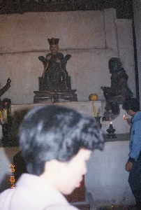 Bối cảnh là không gian bên trong một ngôi đền, chùa hoặc đình làng. Một bàn thờ được đặt sát bức tường cũ kỹ. Trên bàn thờ có nhiều pho tượng thờ bằng gỗ. Tượng chính ở giữa mô tả một vị thần hoặc một nhân vật lịch sử mặc trang phục triều đình, đội mũ quan, ngồi trên ngai. Hai bên là các tượng phụ, trong đó có một tượng mặt xanh có thể là thần hộ pháp.。Một người đàn ông mặc áo khoác màu xanh lam đang thực hiện nghi lễ, dùng bật lửa để thắp một cây nến trên chân đèn dầu. Phía trước bàn thờ có các vật phẩm cúng lễ như một quả tròn màu vàng (giống quả bưởi) và hương (nhang) cắm trong bình. Ở phía tiền cảnh, hình ảnh bị che khuất một phần bởi đầu của một người đang đứng xem.。Trên ảnh có dấu thời gian của máy ảnh màu cam ghi '20 10:40'.。（日本語訳）。背景は、寺院、仏塔、あるいは村の集会所の内部空間です。一つの祭壇が、古びた壁際に置かれています。祭壇の上には、多くの木製の祭祀像があります。中央の主神像は、朝廷の衣装を身に着け、官吏の帽子をかぶり、玉座に座っている神または歴史上の人物を描写しています。両脇には脇侍像があり、その中には護法神と思われる青い顔の像が一体あります。青い上着を着た男性が儀式を行っており、ライターを使って油灯の燭台にあるろうそくに火を灯しています。祭壇の前には、黄色い丸い果物（ザボンのような）や、香炉に立てられた線香（お香）といった供物があります。前景では、見物している人の頭によって、画像が一部隠されています。写真には、「20 10:40」と記されたオレンジ色のカメラのタイムスタンプがあります。