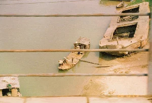 Bức ảnh được chụp từ trên cao, có thể là từ một cây cầu, nhìn xuống một con sông hoặc kênh. Ở giữa ảnh là một chiếc ghe gỗ có mái che tạm. Trên ghe có vài người đang hoạt động; một người ở đuôi ghe, và một người khác đội nón lá đang ở trong khoang có mái che. Họ dường như đang chuẩn bị cập vào bờ sông đầy cát. Bên phải, một chiếc sà lan lớn bằng bê tông hoặc kim loại, trông cũ kỹ và có thể đã bị bỏ hoang, đang nằm trên bờ. Dựa vào sà lan có một vài bánh xe đạp. Ở phía xa hơn cũng có một chiếc thuyền nhỏ khác. Khung cảnh cho thấy hoạt động sinh hoạt và vận tải trên sông. Ở góc dưới của ảnh có dấu ngày tháng màu cam '20 02 '94'.。（日本語訳）。写真は上から、おそらく橋の上から、川か運河を見下ろして撮影されたものです。小舟の上では数人が活動しており、一人は船尾に、もう一人はノンラー（菅笠）をかぶって屋根のある船室にいます。彼らは砂浜の岸に着岸する準備をしているようです。右側には、コンクリートか金属でできた大きなはしけが岸にあり、古びていて、おそらく放置されているように見えます。はしけには数台の自転車が立てかけられています。さらに遠くにも、もう一艘の小さな船があります。この光景は、川での生活や輸送活動を示しています。写真の下隅には、オレンジ色で「'20 02 '94」という日付のスタンプがあります。