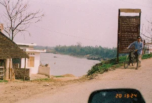 Bức ảnh chụp một khung cảnh ven sông ở miền Bắc Việt Nam vào khoảng năm 1994. Địa điểm là một con đường đất nằm cạnh một con sông rộng, có khả năng là khu vực gần cầu Việt Trì, tỉnh Phú Thọ.。Bên phải đường có một tấm biển báo lớn màu nâu ghi các quy định giao thông. Nội dung có thể đọc được bao gồm:。"QUI-ĐỊNH。QUA CẦU VIỆT TRÌ。CÁC LOẠI PHƯƠNG TIỆN QUA PHẢI CHẤP HÀNH QUI ĐỊNH。1- CẤM CÁC LOẠI XE TỪ 15 TẤN TRỞ LÊN。2- XE XÍCH PHẢI ĐEO GUỐC VÀO XÍCH HOẶC LÓT VÁN" (Chữ "XE XÍCH" và "LÓT VÁN" được suy đoán từ các ký tự hơi mờ trong ảnh gốc là "XE KÍCH" và "LẤT VẤN")。"3- XE MÁY ĐI CÙNG CHIỀU VỚI Ô-TÔ。4- XE THÔ SƠ ĐI CÙNG CHIỀU VỚI Ô TÔ"。Các dòng chữ còn lại ở phía dưới bị mờ và không thể đọc rõ.。Hoạt động chính trong ảnh là một người đàn ông đang dắt chiếc xe đạp đi lên con dốc trên đường đất. Về các vật thể, bên trái là một ngôi nhà cấp bốn lợp mái ngói và một ngôi nhà xây khác. Dưới sông có một vài chiếc thuyền (tàu) đang neo đậu và một chiếc phao tiêu màu đỏ. Một cái cây trụi lá đứng gần nhà, cho thấy thời điểm chụp là vào mùa đông hoặc đầu xuân. Ở góc dưới bức ảnh, có thể thấy một phần gương chiếu hậu của ô tô với đồng hồ điện tử hiển thị giờ là "20 13:24".。（日本語訳）。この写真は1994年頃にベトナム北部で撮影された川沿いの風景です。場所は広い川の隣にある未舗装の道で、おそらくフート省ヴィエットチー橋の近くの地域です。道の右側には、交通規則が書かれた大きな茶色の看板があります。読み取れる内容は以下の通りです：。「規則。ヴィエットチー橋を渡る際の。通行する各種車両は規則を遵守しなければならない。1- 15トン以上の各種車両は通行禁止。2- キャタピラ車はキャタピラにシューを装着するか、板を敷かなければならない」（「XE XÍCH」と「LÓT VÁN」の文字は、元の写真の少しぼやけた文字「XE KÍCH」と「LẤT VẤN」から推測されたものです）。「3- バイクは自動車と同じ方向に進むこと。4- 軽車両は自動車と同じ方向に進むこと」。下部の残りの文字はぼやけていて、はっきりと読み取れません。写真の中の主な活動は、男性が未舗装の坂道を自転車を押して登っているところです。物体については、左側には瓦屋根の平屋と、もう一軒のレンガ造りの家があります。川には数隻の船（ボート）が停泊しており、赤いブイが一つあります。家の近くには葉の落ちた木が一本立っており、撮影時期が冬か早春であることを示しています。写真の下隅には、自動車のサイドミラーの一部が見え、デジタル時計が「20 13:24」と表示されています。