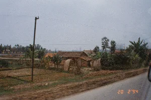 Bức ảnh chụp một khung cảnh nông thôn ở Việt Nam, nhìn từ một phương tiện đang di chuyển trên đường nhựa. Ở góc dưới bên phải của bức ảnh có dấu ngày tháng "20 2'94", cho thấy ảnh được chụp vào ngày 20 tháng 2 năm 1994. Cảnh vật bao gồm những ngôi nhà cấp bốn lợp ngói đỏ, tường gạch mộc hoặc tường đất đặc trưng của vùng nông thôn. Bên cạnh đó là các công trình phụ như lán, chòi có mái vòm được lợp bằng rơm hoặc lá. Xung quanh các ngôi nhà là những thửa ruộng đất đã được thu hoạch hoặc đang chuẩn bị cho vụ mùa mới. Nhiều loại cây cối như cây chuối được trồng xen kẽ. Hệ thống cột điện và dây điện giăng mắc khắp khu vực, cho thấy sự hiện diện của mạng lưới điện. Ở phía xa, có thể thấy một tòa nhà lớn hơn, sơn màu vàng, có kiến trúc giống một trường học hoặc một cơ quan hành chính.。（日本語訳）。この写真はベトナムの田舎の風景を写したもので、舗装された道路を移動中の乗り物から撮影されたものです。写真の右下隅には「20 2'94」という日付のスタンプがあり、写真が1994年2月20日に撮影されたことを示しています。風景には、赤い瓦屋根、むき出しのレンガ壁、または土壁といった、田舎特有の平屋建ての家々が含まれています。その隣には、藁や葉で葺かれたドーム型の屋根を持つ小屋やあばら家などの付属的な建物があります。家々の周りには、収穫が終わった、あるいは新しい作付けの準備をしている畑が広がっています。バナナの木など、さまざまな種類の木々が点在して植えられています。電柱と電線が地域全体に張り巡らされており、電力網の存在を示しています。遠くには、黄色に塗られた、学校か行政機関のような建築様式の、より大きな建物が見えます。