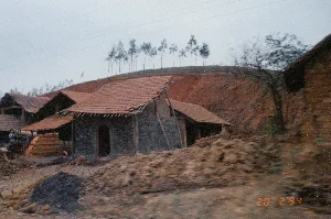 Dấu hiệu duy nhất có thể đọc được là ngày tháng "20 2 '94" ở góc dưới bên phải của ảnh.。Bức ảnh chụp lại khung cảnh một cơ sở sản xuất gạch thủ công, có khả năng nằm ở một vùng trung du của Việt Nam. Trung tâm của bức ảnh là những lò nung gạch (lò gạch) được xây dựng依 sườn một quả đồi đất đỏ. Các lò gạch này có cấu trúc đơn giản với tường gạch thô hoặc trát bùn, mái lợp ngói đỏ đã cũ. Một trong những lò nung có cửa vào hình vòm đặc trưng. Xung quanh khu vực lò là những đống đất sét lớn, là nguyên liệu để sản xuất gạch. Ở phía bên trái, có thể thấy một chồng gạch thành phẩm được xếp ngay ngắn và một vài người đang làm việc. Phía sau, quả đồi bị đào xới để khai thác đất, và trên đỉnh đồi là một hàng cây cao, khẳng khiu. Phía trên có các đường dây điện chạy ngang qua bầu trời. Toàn bộ khung cảnh cho thấy một hoạt động sản xuất công nghiệp nhỏ, mang tính thủ công đặc trưng của thời kỳ đó.。（日本語訳）。写真の右下隅で読み取れる唯一の印は、「94年2月20日」という日付です。この写真は、ベトナムの丘陵地帯にあると思われる、手作りのレンガ製造施設の光景を捉えています。写真の中心には、赤土の丘の斜面に沿って建てられたレンガを焼く窯（レンガ窯）があります。これらのレンガ窯は、粗いレンガまたは泥で塗られた壁と、古くなった赤瓦で葺かれた屋根を持つ、簡単な構造をしています。窯の一つには、特徴的なアーチ型の入口があります。窯の周辺には、レンガ製造の原料である大きな粘土の山があります。左側には、きちんと積み上げられた完成品のレンガの山と、作業をしている数人の姿が見えます。後方では、土を採掘するために丘が掘り起こされており、丘の上には高く痩せ細った木々が並んでいます。上方には、空を横切る電線があります。全体の光景は、その時代に特徴的な、手工業的な性質を持つ小規模な工業生産活動を示しています。