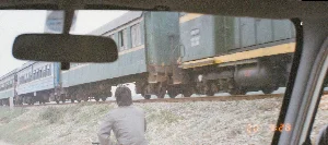 Bức ảnh được chụp từ bên trong một chiếc ô tô, nhìn ra ngoài qua cửa sổ. Khung cảnh cho thấy một đoàn tàu hỏa đang chạy trên đường ray được đắp cao bằng sỏi đá ở một vùng nông thôn.。Về chữ viết, trên thân đầu máy xe lửa màu xanh lá cây có thể đọc được dòng chữ và số "D13E - 710". Ngoài ra, có một dấu thời gian kỹ thuật số màu cam ở góc dưới bên phải hiển thị "20 9:28". Một số chữ khác trên toa tàu và trên một miếng dán phản chiếu trong gương chiếu hậu nhưng không thể đọc rõ.。Hoạt động chính đang diễn ra là đoàn tàu khách đang di chuyển. Đoàn tàu bao gồm một đầu máy diesel màu xanh lá cây sọc vàng và nhiều toa hành khách. Các toa xe có hai màu sắc khác nhau: một số toa màu xanh lá cây và kem, một số toa khác ở phía xa hơn có màu xanh dương và trắng. Bên dưới đường ray, một người đang ngồi trên xe đạp, quay lưng về phía máy ảnh, dường như đang dừng lại để xem tàu đi qua. Các vật thể nổi bật trong ảnh bao gồm đoàn tàu hỏa (tàu), đường ray, một người đi xe đạp, và một phần nội thất của chiếc ô tô như gương chiếu hậu và khung cửa sổ.。（日本語訳）。写真は車内から窓越しに外を向いて撮影されたものです。風景は、田舎の砂利で高く盛られた線路の上を走っている列車を示しています。文字については、緑色の機関車の車体には「D13E - 710」という文字と数字が読み取れます。また、右下隅には「20 9:28」と表示されたオレンジ色のデジタルタイムスタンプがあります。行われている主な活動は、旅客列車が移動していることです。列車は、黄色い縞模様のある緑色のディーゼル機関車1両と、多くの客車で構成されています。車両には2つの異なる色があります。一部の車両は緑色とクリーム色で、さらに遠くにある他の車両は青色と白色です。線路の下では、一人の人が自転車に座り、カメラに背を向けて、列車が通り過ぎるのを見るために立ち止まっているようです。写真の中で目立っている物体には、列車、線路、自転車に乗った人、そしてバックミラーや窓枠といった車内の一部が含まれます。