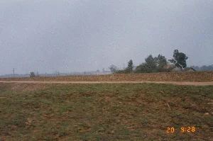 Bức ảnh chụp một cảnh quan nông thôn đặc trưng của Việt Nam, có khả năng là ở vùng trung du. Nổi bật trong ảnh là một con đê đất lớn chạy dài theo chiều ngang. Trên mặt đê là một con đường đất. Phía trước con đê là một triền cỏ, hình ảnh hơi nhòe cho thấy có thể được chụp từ một phương tiện đang di chuyển. Phía sau con đê là một vùng đất bằng phẳng, có thể là cánh đồng. Ở phía xa, có thể nhìn thấy những hàng cột điện, cây cối và một vài ngôi nhà mái ngói của người dân. Bầu trời trong ảnh có màu xám và u ám. Ở góc dưới bên phải có dấu thời gian kỹ thuật số màu cam ghi '20 9:28'.。（日本語訳）。この写真はベトナムの典型的な田園風景を写しており、おそらく中遊地域のものでしょう。写真の中で目立っているのは、水平に長く伸びる大きな土手です。土手の上には未舗装の道があります。土手の手前には草の斜面があり、画像が少しぼやけていることから、移動中の乗り物から撮影された可能性が示唆されます。土手の後ろには平坦な土地が広がっており、おそらく田畑でしょう。遠くには、電柱の列、木々、そして住民の瓦屋根の家がいくつか見えます。写真の空は灰色でどんよりとしています。右下隅には、「20 9:28」と記されたオレンジ色のデジタルタイムスタンプがあります。